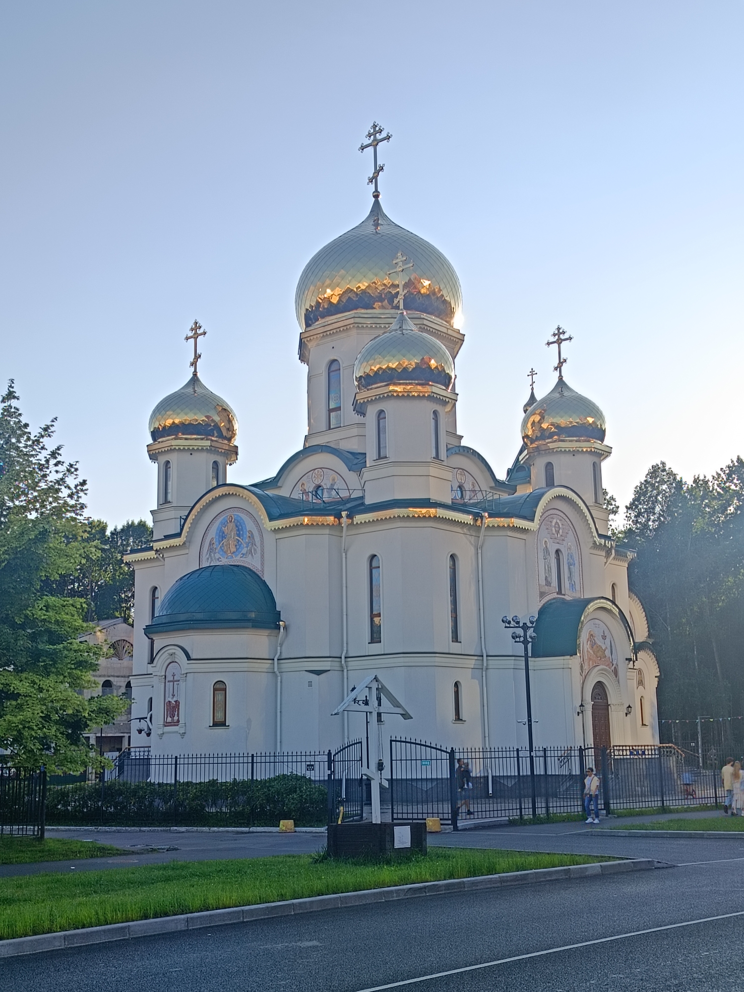 The image shows a large, white, Orthodox church with multiple golden domes topped with crosses, set against a clear, pale blue sky. The church has a grand facade with decorative elements and religious imagery in mosaics. There is a fence in front of the church, with a black wrought-iron design. Beyond the fence, there are trees and greenery. Several people are visible near the fence and on the walkway in front of the church, appearing to be visitors or parishioners. The foreground shows a paved road and a grassy area. This building is the Church of the Resurrection of Christ, located in the city of Saint Petersburg, Russia.