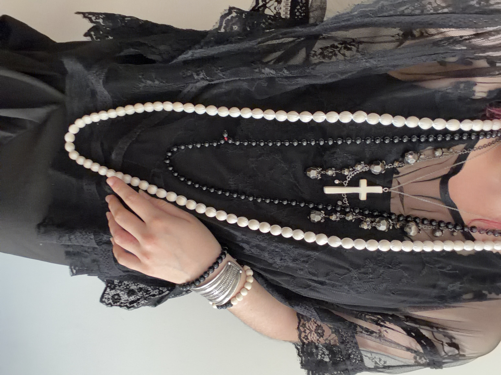 The image shows a close-up of a person wearing multiple necklaces and bracelets, and a sheer black lace top. The necklaces are layered, with one long strand of large white beads, a strand of smaller black beads with dark grey pendants, and another strand with a white cross pendant. The bracelets on the person's wrist consist of a thick silver cuff adorned with intricate patterns and two beaded bracelets, one with black beads and another with white beads. The person's hand is visible, resting on their chest, and they are wearing a black garment underneath the lace top. The background is a plain, light-colored wall. There is no discernible information or context to suggest a location in the city of Chelyabinsk, Russia.