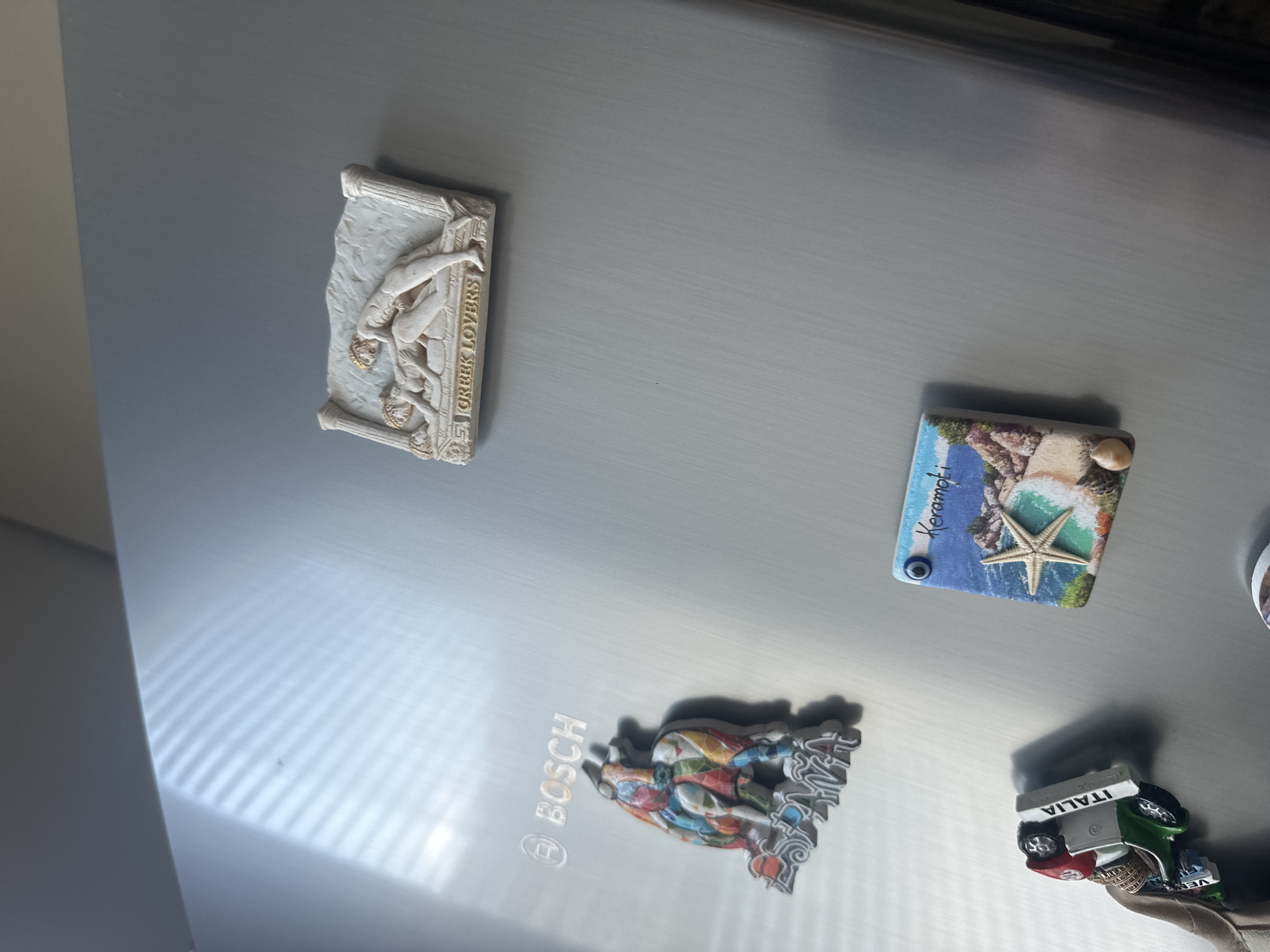 The image is a close-up shot of a collection of magnets on a refrigerator door. The refrigerator is a silver Bosch model. Among the magnets, there is a 3D magnet depicting a scene with two figures, labeled "GREEK LOVERS." Another magnet features a colorful bull with the word "ESPAÑA" below it, and a Bosch logo is visible above it. There is also a magnet shaped like a small car with the word "ITALIA" on its side, and a square magnet with a beach scene labeled "Keramoti" and a blue evil eye charm. The lighting suggests the sun is shining through blinds, casting striped shadows across the refrigerator door. The image was taken in Ruse, Bulgaria.