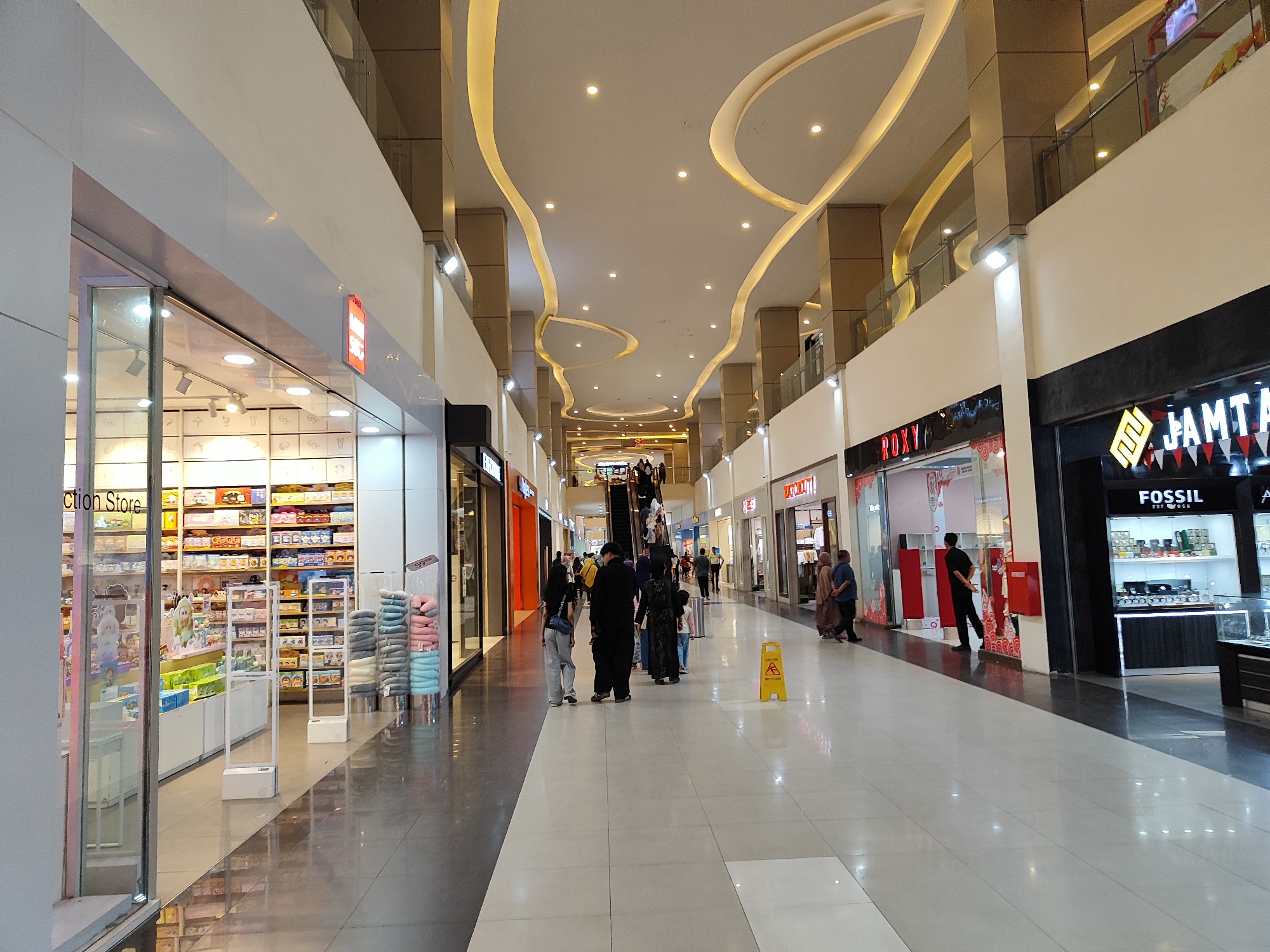 The image depicts the interior of a modern shopping mall, likely the Roxy Square mall located in Sempusari, Indonesia, based on the context provided. The mall features a spacious, well-lit corridor with a polished floor reflecting the ambient light from the ceiling and shop displays. The ceiling is adorned with decorative, curvilinear lighting fixtures and recessed spotlights. Along both sides of the corridor are various retail stores.
On the left side, a store labeled "Collection Store" is visible, featuring a glass facade displaying merchandise. Further down, more shops are present with visible signage such as "ROXY" and "FOSSIL". The stores exhibit a variety of products, from consumer goods to accessories.
Several individuals are seen walking through the mall. In the foreground, a few people are walking towards the viewer, while others are moving away. In the mid-ground, a group of people are gathered near an escalator, some ascending. A yellow caution sign warning of a wet floor is placed on the right side of the corridor. The overall atmosphere is that of a busy commercial environment.