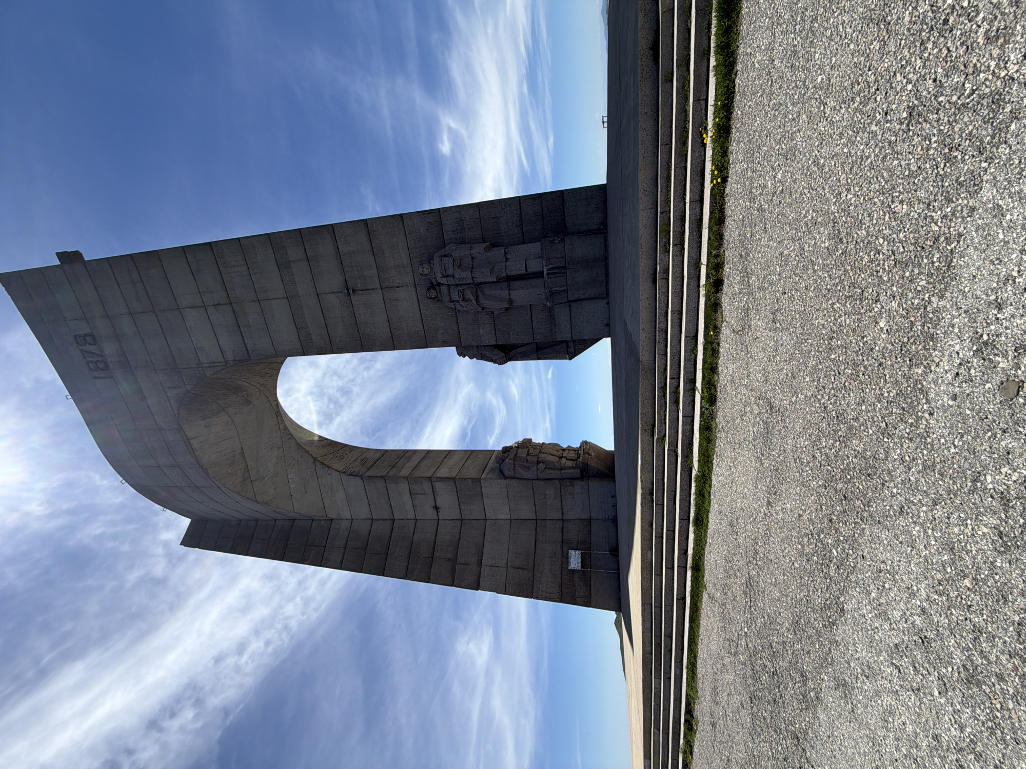 A ground-level, wide-angle shot shows the Shipka Monument, a large concrete structure, against a blue sky with wispy clouds. The monument features a large arch shape with a concrete roadway leading to it. The number "1878" is visible on the left side of the arch. On the top of the arch are relief sculptures of figures. To the right of the monument, a set of concrete stairs leads up, with some grass and small yellow flowers growing along the edges of the stairs. A grey, rough concrete walkway fills the foreground. The photo is rotated counter-clockwise. The monument is located in Shipka, near the town of Beli Osam, Bulgaria.