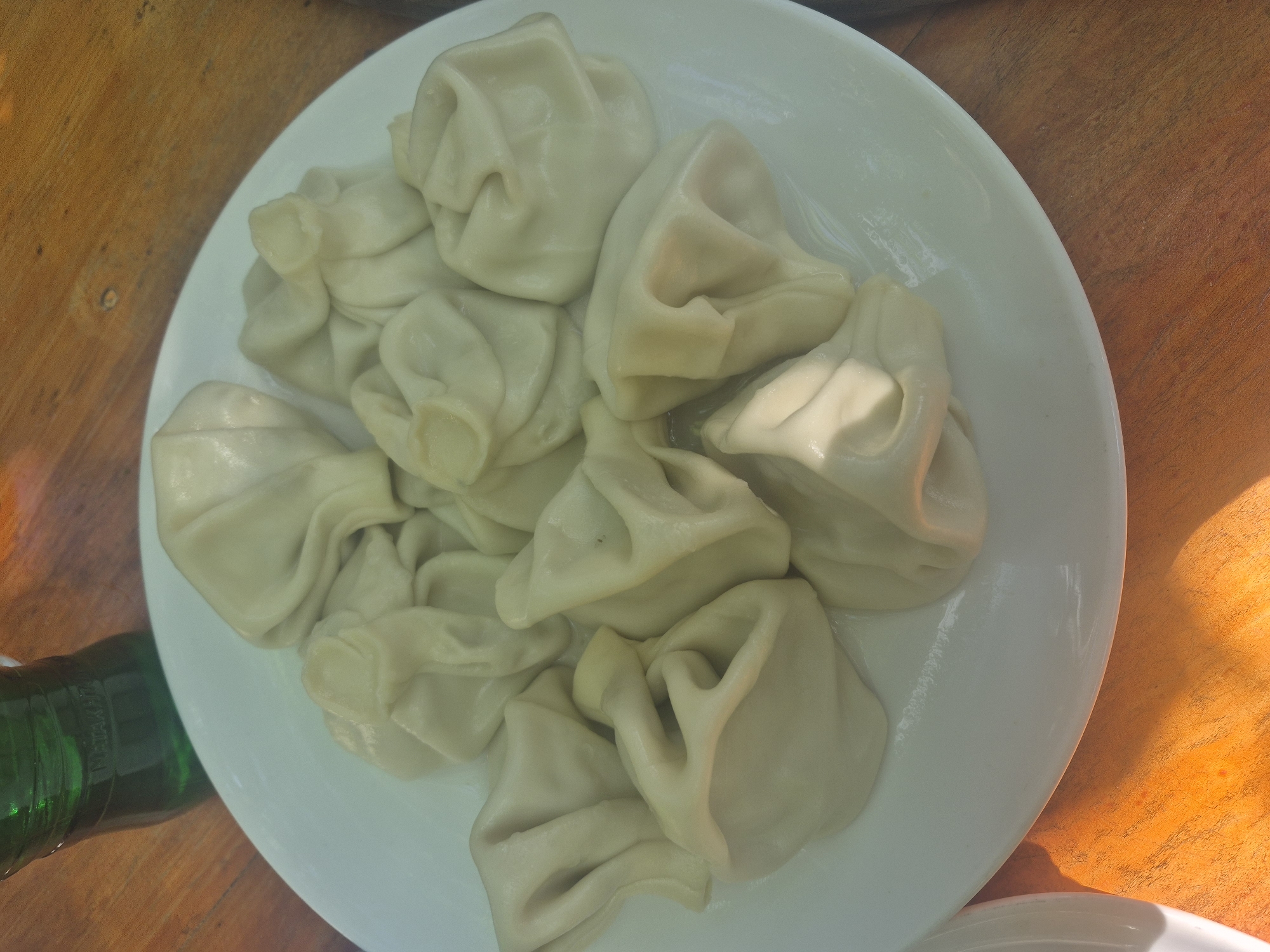 A top-down shot shows a white plate filled with approximately a dozen Khinkali (Georgian dumplings) resting on a wooden surface, possibly a table. A dark green bottle is partially visible on the lower left. The light suggests an outdoor or well-lit environment. No people are in the view. No location information is available.