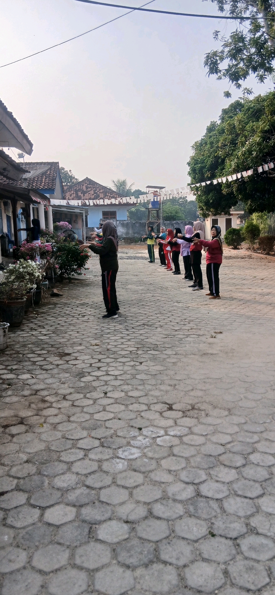 In the city of Metro, Indonesia, a group of women are performing outdoor exercises. The scene is set in what appears to be a schoolyard or community space, paved with hexagonal interlocking concrete blocks. To the left, a low-slung building with a tiled roof and open-air covered walkways is visible. Potted flowering plants, predominantly pink and white, line the walkway in front of the building.
In the center and extending to the right, approximately eight women, all wearing hijabs and athletic attire, are standing in a line, facing to the right. They are engaged in a synchronized physical activity, with their arms extended forward and slightly upward, palms facing each other. Their movements suggest a coordinated exercise routine. The women are dressed in various colors, including red, pink, light purple, and dark green.
Behind the women and the building, trees with dense green foliage are present. A white string of small flags or banners is strung across the upper right portion of the image, above the trees, adding a decorative element to the outdoor setting. The sky is overcast, and the overall lighting suggests it is either early morning or late afternoon. In the background, a raised platform structure, possibly for observation or a water tank, is visible.