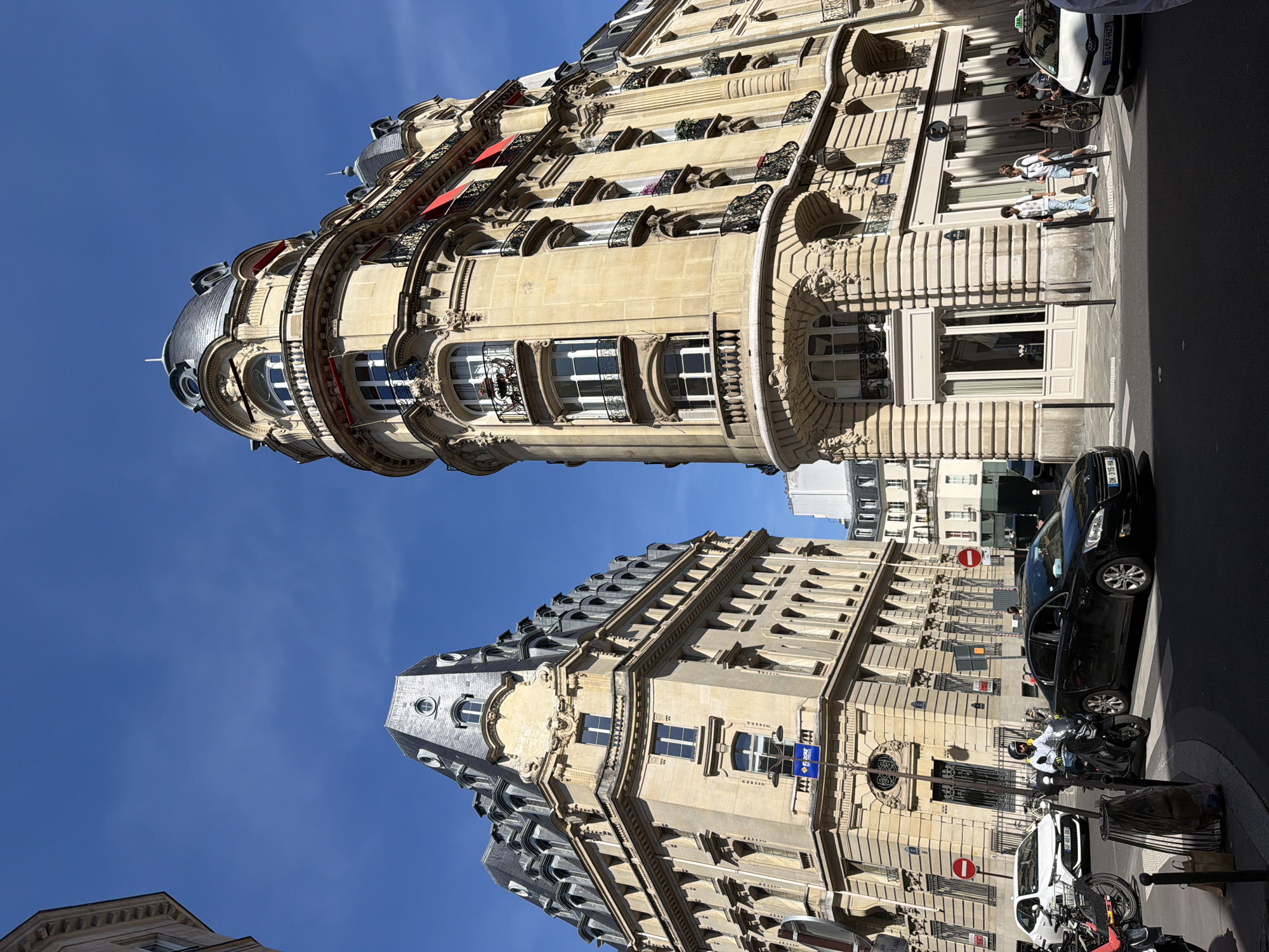 The image shows two ornately designed buildings in Paris, France. The first building is rounded with several balconies, and the second building has a gray roof. The sky is clear and blue. Along the street, several people are walking, and vehicles such as cars, scooters, and bicycles are parked. There is a street sign nearby.