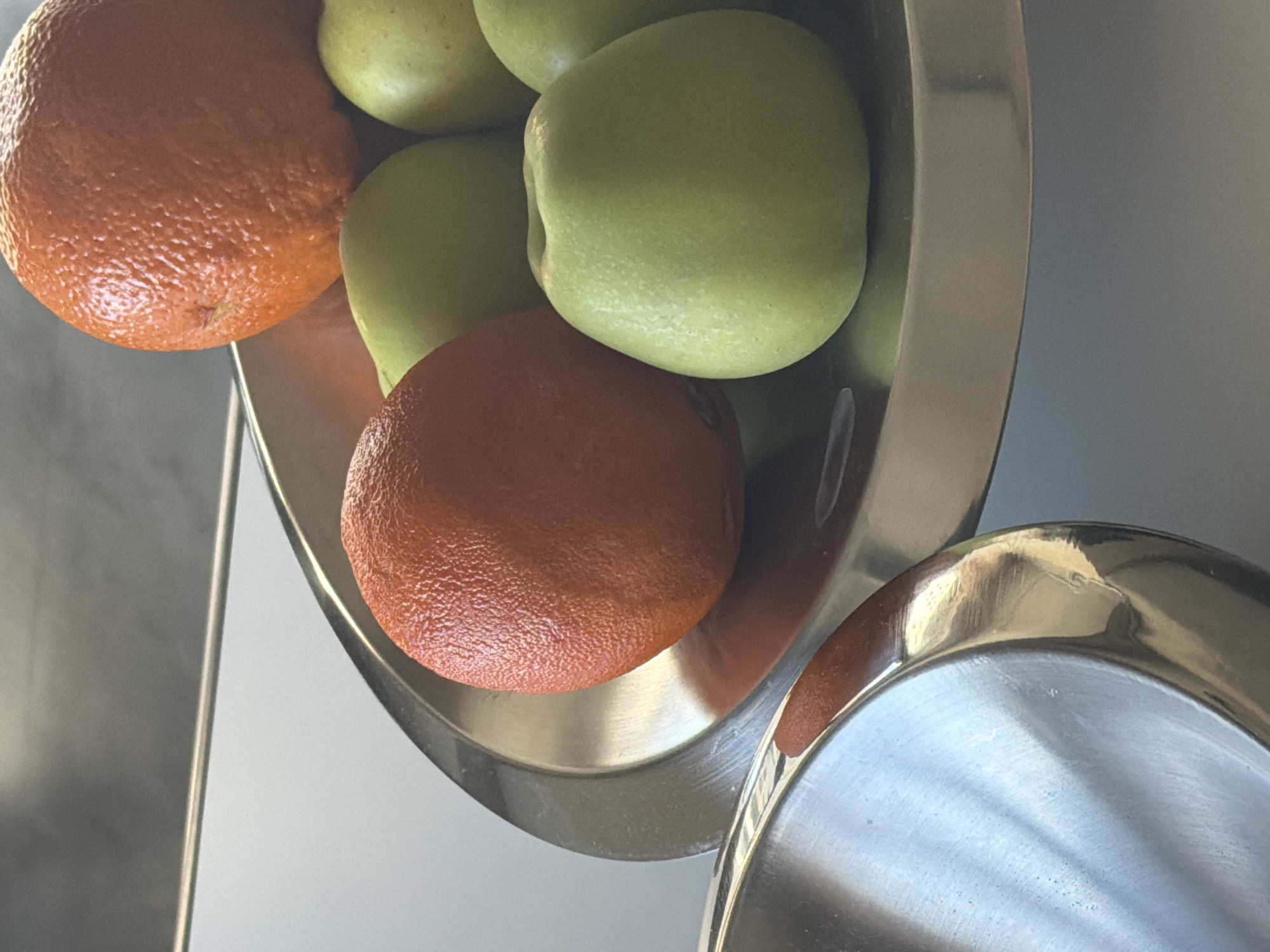 A close-up shot depicts a stainless steel bowl containing a mixture of oranges and green apples. The bowl is positioned at an angle, with some fruit appearing to spill out. The background is a blurred, light gray surface, possibly a kitchen counter or wall. The scene suggests a domestic kitchen setting. There is no indication of the location being Yerevan, Armenia, or any people or specific actions taking place.
