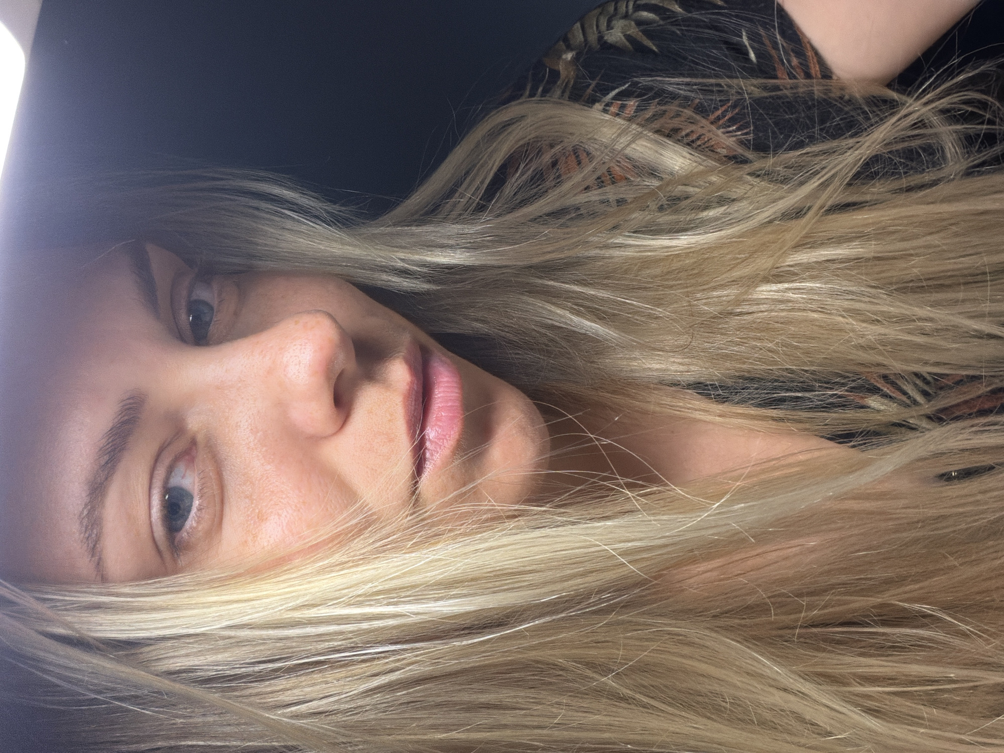 A close-up, overhead shot features a young Caucasian woman with blonde hair and blue eyes. She is looking directly into the camera, with a slight smirk. Her hair is long and layered, with some strands falling across her face. She is wearing a black shirt with a pattern of orange and green leaves. The background is dark and out of focus. There is no visible indication of the location being Votkinsk, Russia.