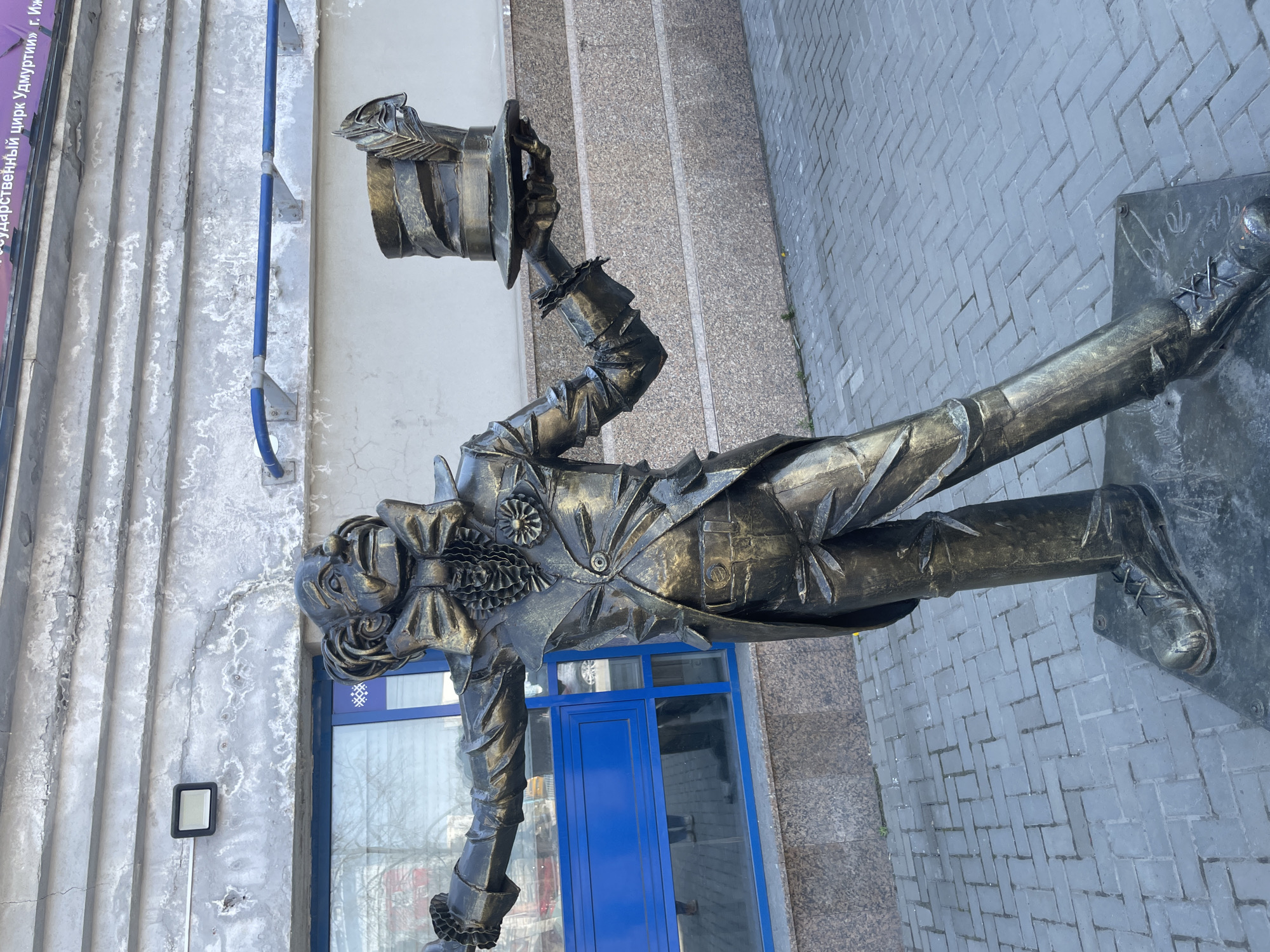 The image shows a bronze statue of a clown, seemingly tipping his hat, located outside the State Circus of Udmurtia in Izhevsk, Russia. The statue stands on a paved area and is placed in front of a building with a blue door and window. The building has a weathered, light-colored facade. A blue pipe runs along the wall near the statue. Steps are also visible in the background. The statue is detailed, with elaborate clothing and features.