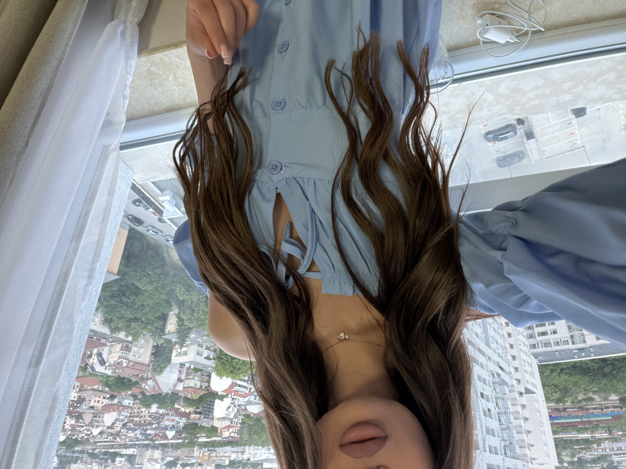 A woman is depicted from the upper torso, with her face cropped above the mouth, showcasing her long, wavy brown hair, manicured nails, and light blue off-shoulder dress featuring front buttons and a tied neckline. A delicate gold necklace with a small, light-colored charm is visible around her neck. She is positioned indoors, adjacent to a window that provides an elevated view of the city of Plovdiv, Bulgaria.
The environment outside the window reveals a dense urban landscape. In the immediate background, a modern, tall, white building with a distinct grid pattern dominates the view on the left. Beyond this, the cityscape extends with numerous multi-story residential buildings, many characterized by traditional red-tiled roofs, interspersed with mature trees and green spaces. A ground-level parking area is visible directly below the window, containing several parked cars. The windowsill, a light-colored surface, holds a white charging adapter with a coiled cable. A white sheer curtain hangs on the right side of the window frame.