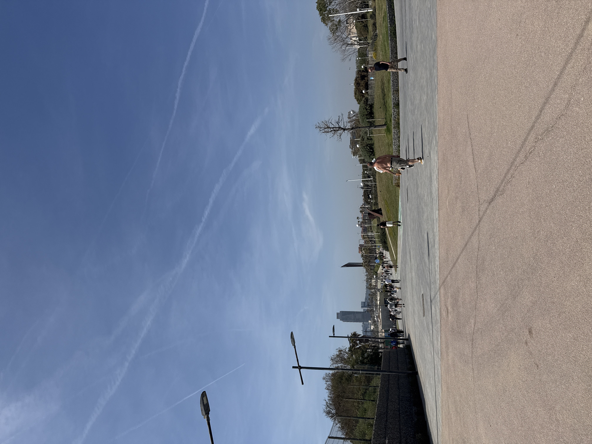 The image shows a wide pedestrian walkway near the city of Barcelona, Spain.
**Environment:** A clear blue sky with contrails is visible. The walkway is divided into two distinct sections: a gray paved area and a broader pinkish-tan paved area. A low fence borders the walkway. Lampposts line the walkway. Trees and buildings are visible in the background, including recognizable Barcelona skyline features like the Torre Agbar and the Hotel Arts.
**People and Actions:** Several people are walking along the walkway. A shirtless person in shorts appears to be throwing something. Another person is jogging. In the distance, a large group of people are walking together.