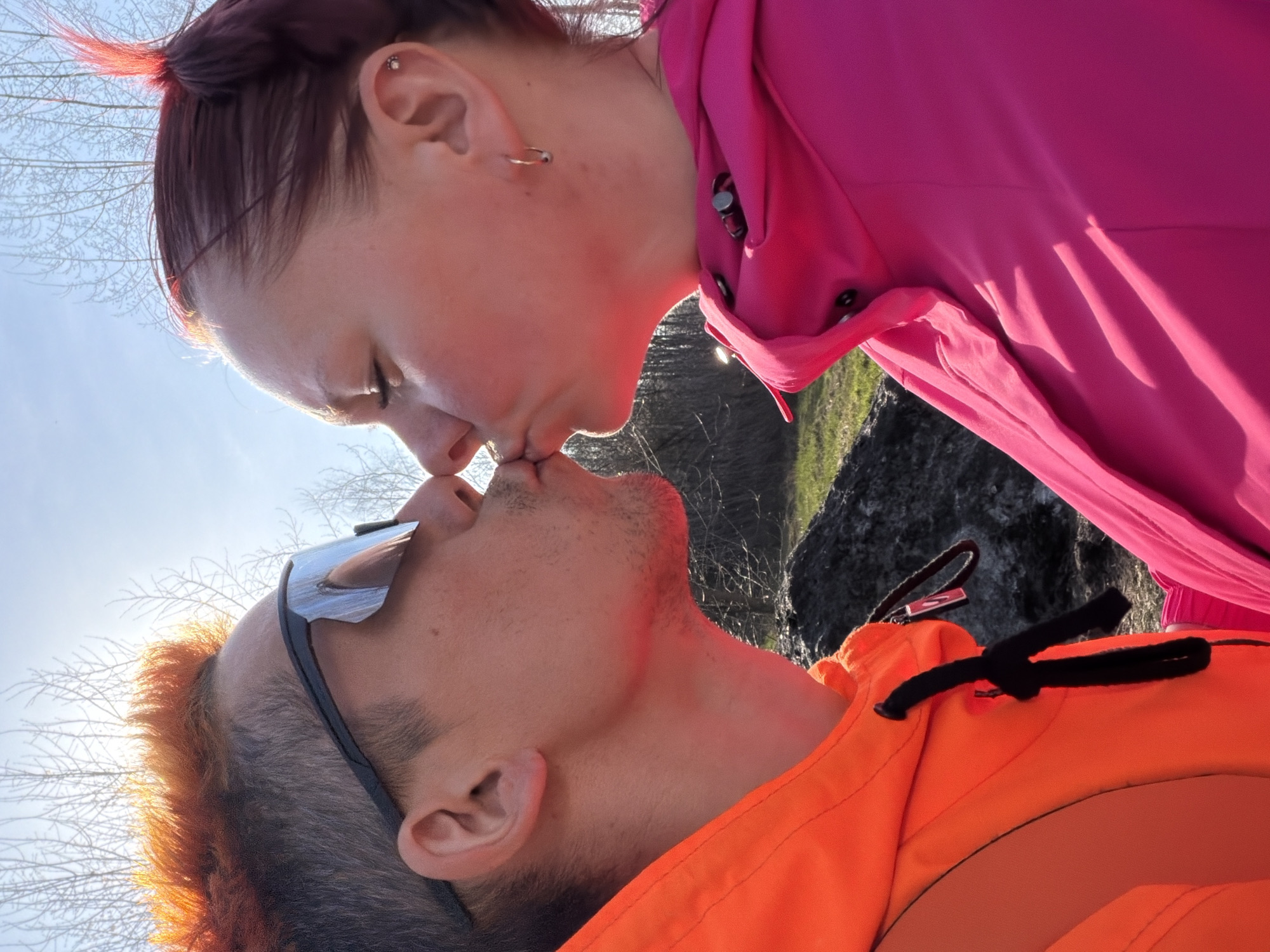 A close-up shot shows a woman with reddish-auburn hair pulled back and wearing a pink buttoned shirt kissing a man with orange and grey hair, sunglasses, and an orange jacket. The background includes a light blue sky, thin bare branches, and a dark rocky area with some greenery. The location is presumed to be in the city of Chaikovsky, Russia.