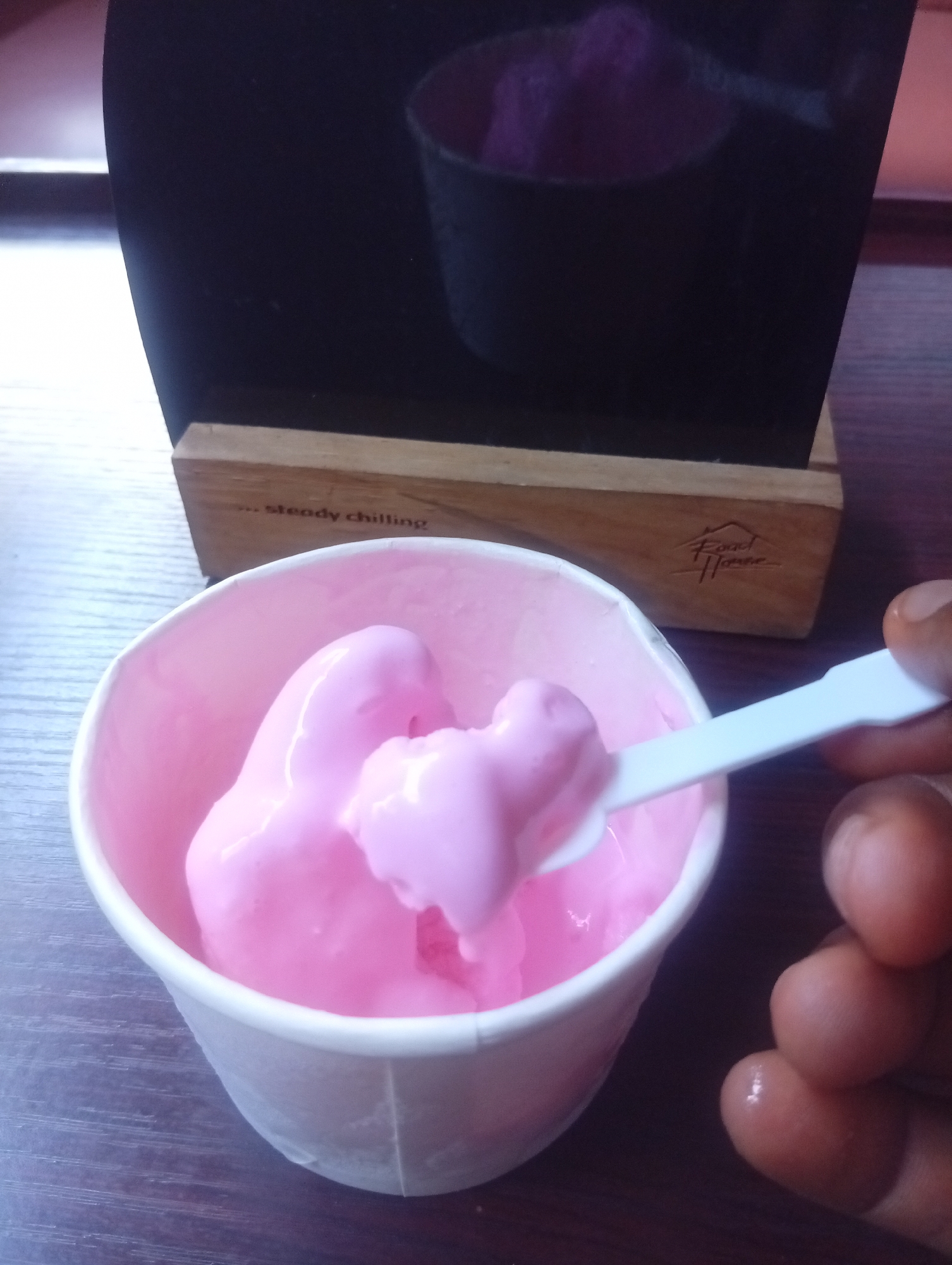A close-up shot focuses on a person about to eat bright pink ice cream from a white paper cup. A hand with dark skin holds a white plastic spoon, which is laden with a scoop of the melting pink ice cream, hovering above the cup. The cup is filled with the same vibrant pink ice cream and rests on a dark, possibly wooden, table surface. In the background, a wooden block serves as a stand for a black, reflective panel. Text is visible on the wooden block, reading "... steady chilling" on the left, and a logo above the words "Food House" on the right. The black panel behind the block shows a blurry reflection of a purple or pink object, potentially another ice cream serving. The scene is set indoors, likely at a dessert shop or cafe, and the overall impression is one of enjoying a sweet, cold treat. The location context provided is Howayi, Nigeria.