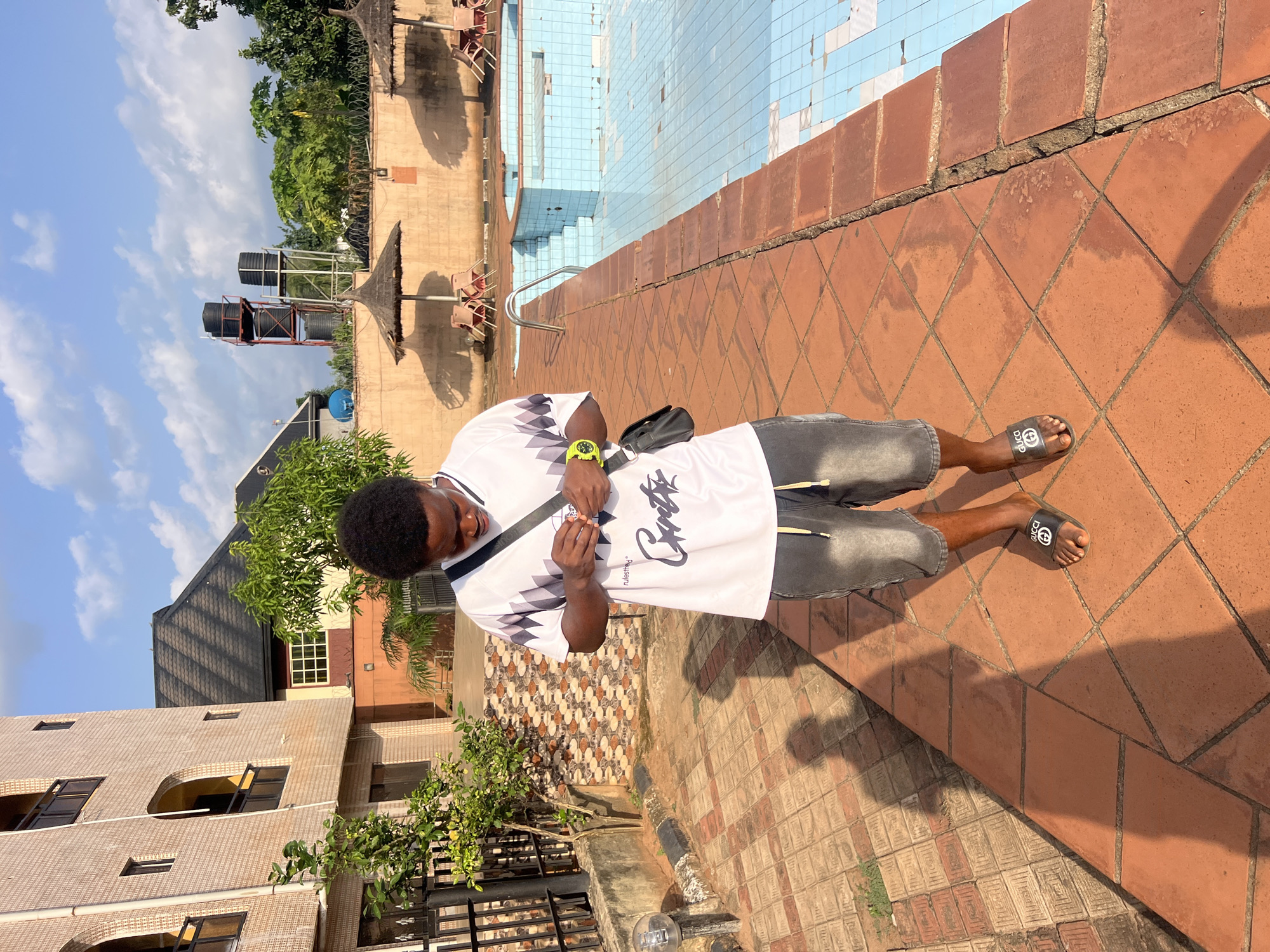 The image displays a person standing outdoors on a bright, sunny day in Nkpor, Nigeria, captured from a rotated perspective. The subject, a male, is dressed in a white short-sleeved shirt featuring black and grey geometric patterns, grey shorts, and black flip-flops. He wears a black crossbody bag and a bright yellow watch on his left wrist. He is depicted with his head bowed, seemingly looking down at his hands.
The setting is a patio paved with reddish-brown square tiles, adjacent to a swimming pool. The pool, tiled in blue and white, appears to be empty or have a very low water level, with some tiles discolored or missing. A metal handrail is visible extending into the pool area.
In the background, under a clear blue sky with sparse white clouds, are two large black water tanks mounted on metal stands. Various buildings are also visible, including one with a light-colored brick facade and others with dark roofs. Lush green trees and vegetation are abundant throughout the scene. Several thatched structures, possibly umbrellas or huts, are located near the pool. Strong shadows cast on the ground indicate bright sunlight, suggesting it is likely mid-day or early afternoon.
Visible text includes the "GUCCI" logo on the black flip-flops. On the person's shirt, the word "rulesthrd" is printed, alongside a larger, stylized script that resembles "Carettas".