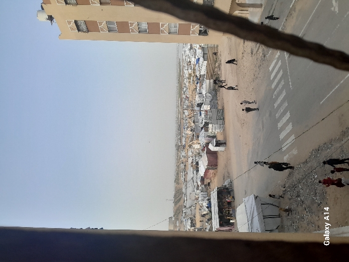 An elevated, downward-angled view captures a scene in Khan Yunis, Palestinian Territory. The image is oriented vertically, depicting a landscape from a height. In the upper left quadrant, a multi-story, light-brown building with multiple rectangular windows is partially visible against a pale blue sky. A dark, out-of-focus horizontal bar spans the lower portion of the image, indicating the viewpoint is from an elevated structure or balcony. Below the building and extending across the mid-ground is a paved road featuring a white pedestrian crosswalk. To the left of the road, and stretching into the background, is an extensive, densely packed encampment consisting of numerous makeshift shelters, primarily constructed from tarpaulins and other light-colored materials. The ground in the encampment and along the unpaved shoulder of the road is dirt or sand, with scattered debris.
Several individuals are present: two figures walk near the crosswalk on the road, two additional figures are positioned further right on the road, and other individuals are visible near the road's edge and within the encampment area. Actions include walking and standing. Overhead, utility wires traverse the scene above the road. The distant horizon appears hazy. A 'Galaxy A14' watermark is present in the bottom right corner of the image.