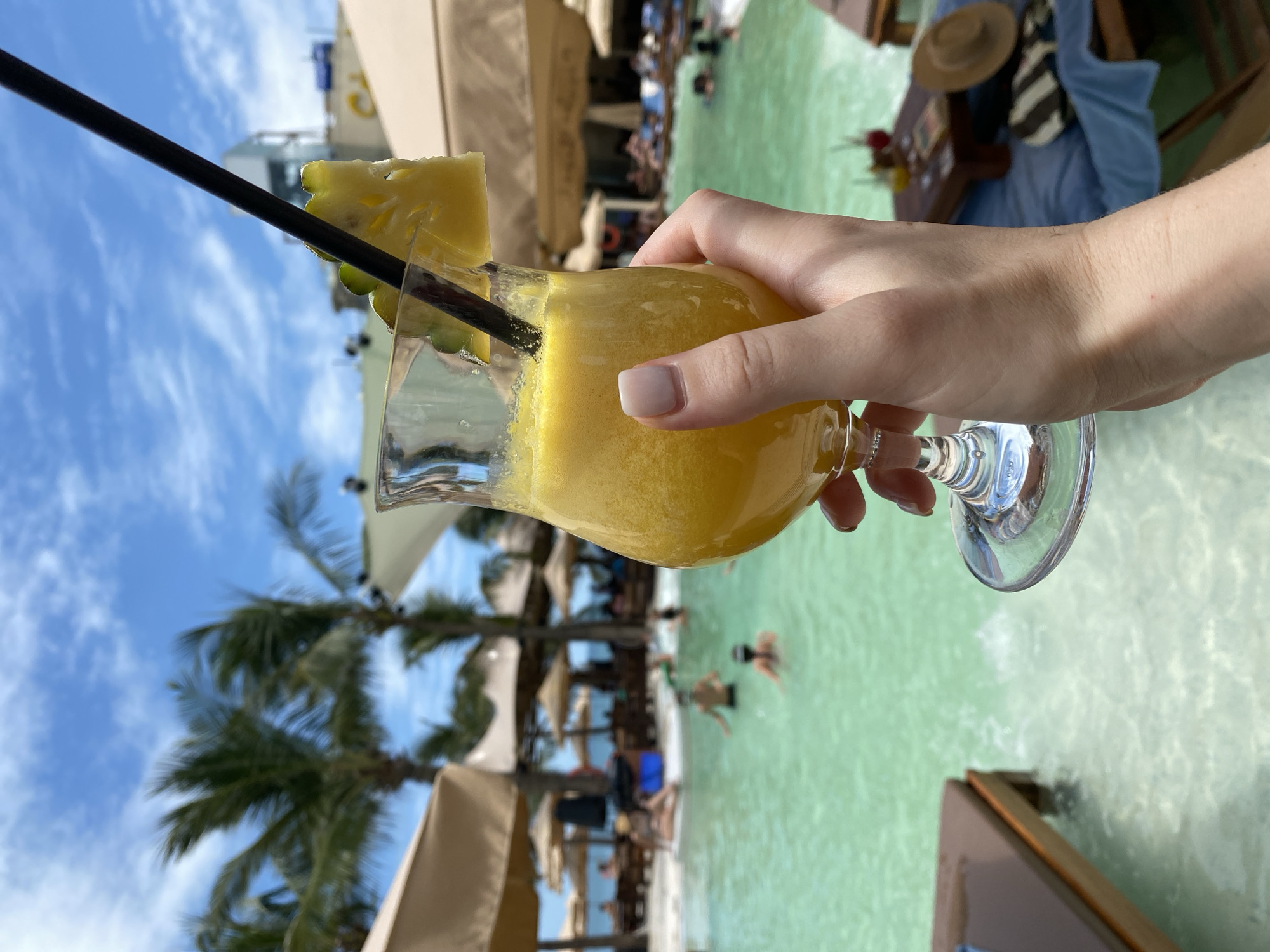 A hand holds a glass of yellow beverage with a slice of pineapple and a straw, in front of a swimming pool. Palm trees and umbrellas are visible in the background, along with several people swimming in the pool. The scene suggests a resort or beach club setting in Da Nang, Vietnam.