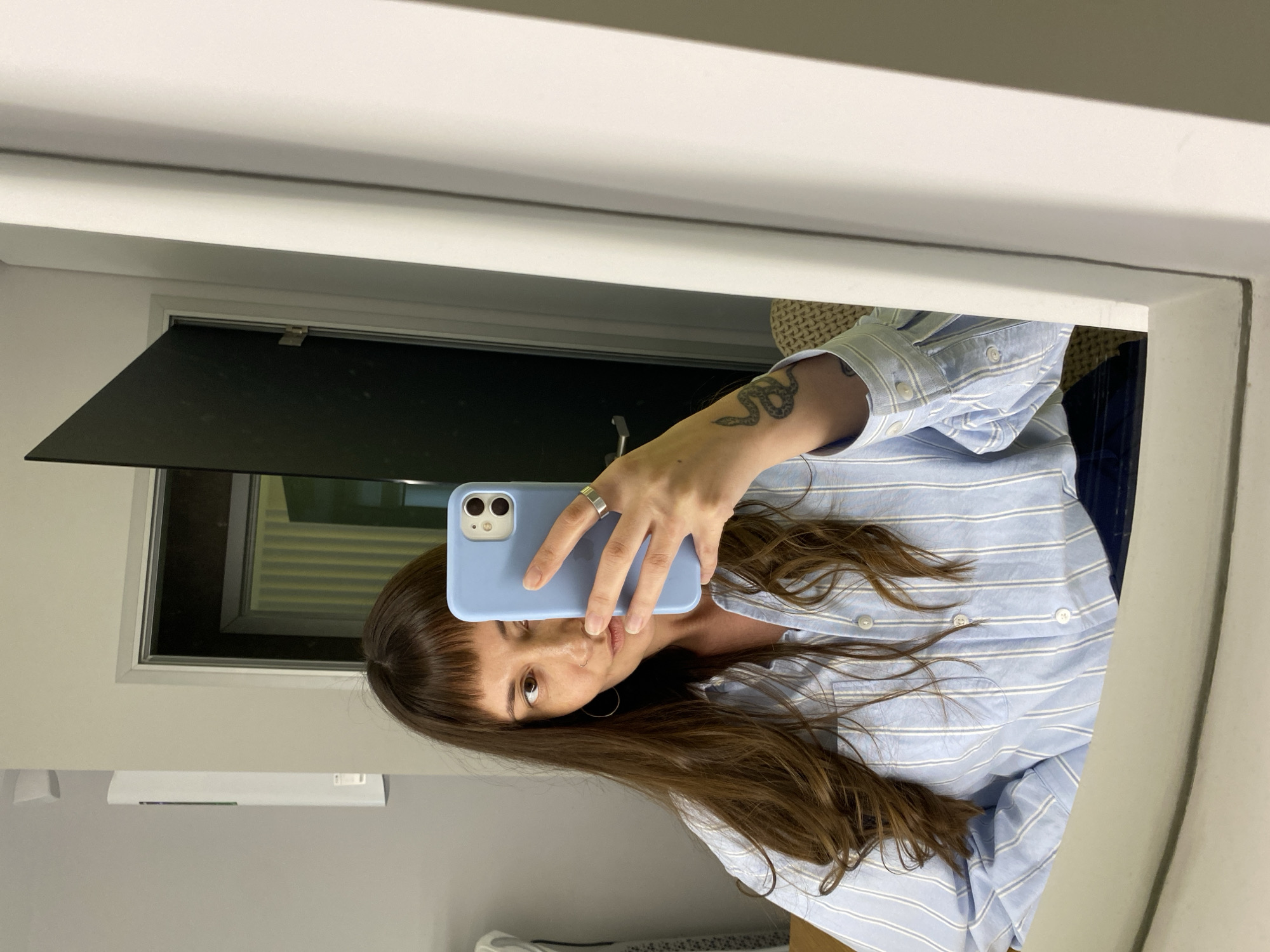 A woman with long brown hair and bangs is taking a selfie in a mirror. She is wearing a light blue and white striped button-up shirt. She is holding a light blue phone in her right hand, which is visible in the mirror. A silver ring is on her right ring finger. She has a snake tattoo on her left forearm, which is also visible. She is wearing a hoop earring in her left ear. The background shows a doorway with a black door, a white wall, and a white cabinet. The scene appears to be indoors, possibly in a hotel room or apartment. The image is a mirror reflection, so it is difficult to determine the exact location. There is no information to suggest this image is from Votkinsk, Russia.