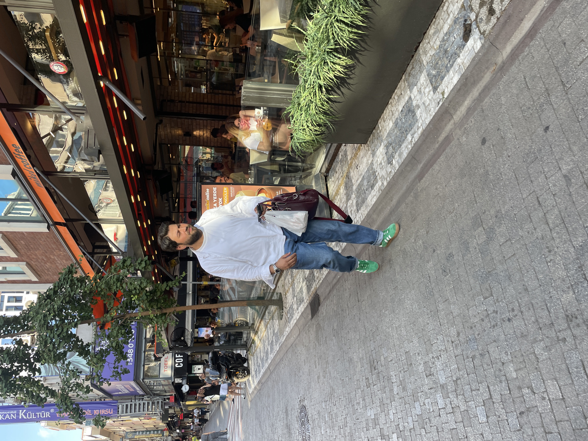 A man with a beard, wearing a white long-sleeved shirt and blue jeans, walks on a cobblestone sidewalk in Kadıköy, Turkey. He is carrying a dark red bag and a white plastic bag. He is wearing green sneakers. The man is walking towards the right side of the frame. Behind him, there is a row of shops and cafes with outdoor seating. A tree with green leaves is growing on the sidewalk. Several people are seated at tables in the cafes, and others are walking on the sidewalk in the background. The environment appears to be a busy urban street.