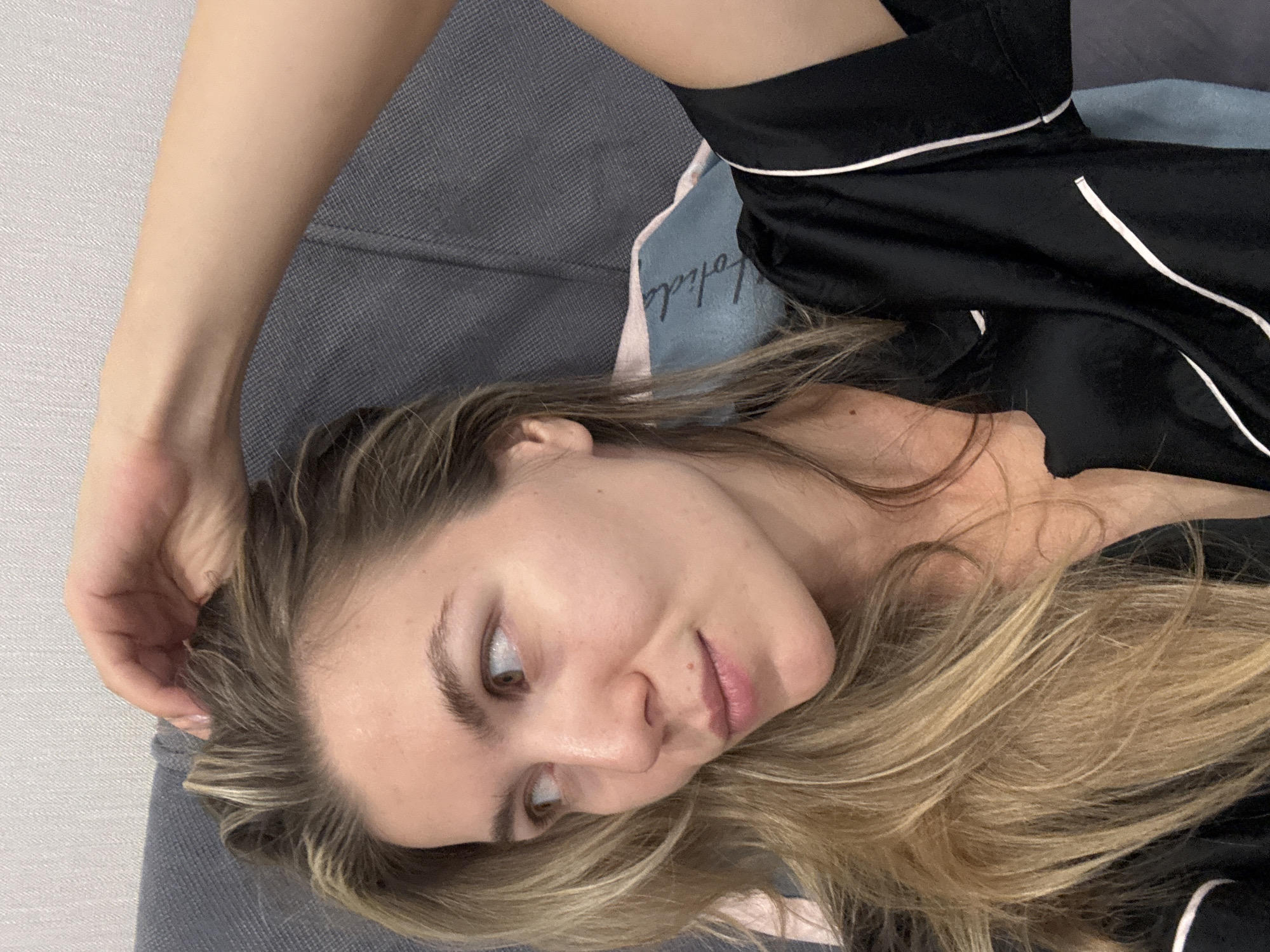 A blonde woman is lying down, her head propped up by a gray pillow and a blue pillow with white lettering on it. Her right arm is raised, with her hand resting on her head, touching her hair. She is wearing a black satin pajama top with white trim. The background appears to be a wall with a textured surface.