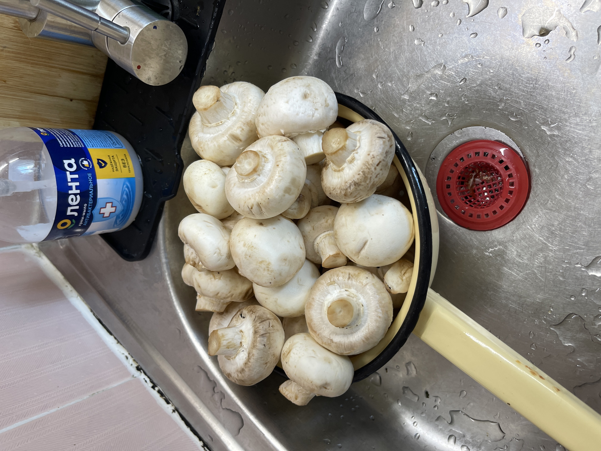 In a stainless steel sink in Khimki, Russia, a colander filled with white button mushrooms sits on the edge. The sink is wet, with water droplets visible on the surface. Next to the sink, a bottle of Lenta antibacterial hand soap is placed on a black mat. A chrome faucet is also visible to the right of the soap bottle. The background shows a tiled wall with a pink herringbone pattern. A red drain cover is in the sink.
