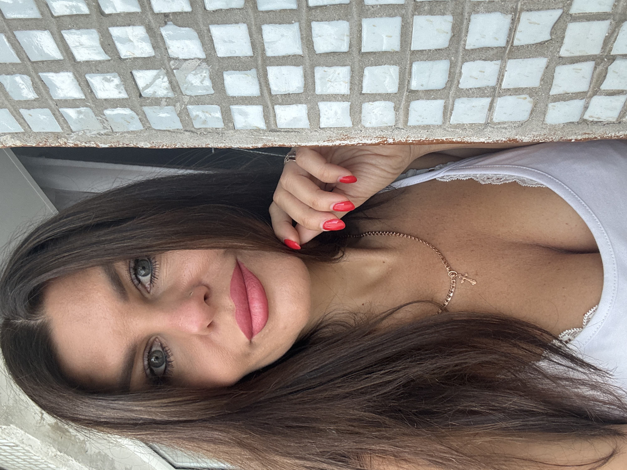 A woman with long, dark brown hair and blue eyes is shown in a close-up portrait. She is wearing a white tank top with lace trim and a rose gold necklace with a cross pendant. Her fingernails are painted bright red. She is looking towards the camera with her left hand resting near her face. The background consists of a tiled wall with white square tiles. The image does not provide any information about a specific location in Chaikovsky, Russia.