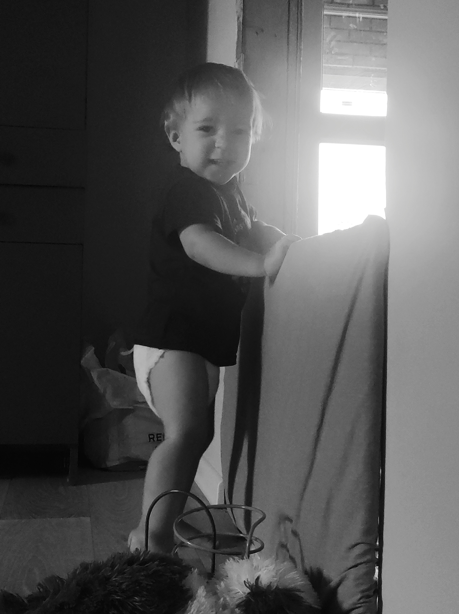 An indoor, black and white photograph captured in Фоки, Russia, depicts a young child, approximately a toddler, standing in an upright position. The child is positioned centrally, facing the viewer, and appears to be smiling. They wear a dark, short-sleeved t-shirt and a light-colored diaper. Both hands are resting on the upper edge of a large, fabric-covered object, likely a piece of furniture such as a crib, playpen, or sofa, which extends along the right side of the frame. To the child's left, a dark wall or door with subtle paneling is visible. To the child's right and behind the furniture, a bright window emits strong light, causing overexposure in that area. The floor is visible in the lower portion of the image, appearing to be made of wood. On the floor in the mid-left, a light-colored bag with the partial inscription "REI" is present alongside other indistinct items. In the immediate foreground, dark, fuzzy objects, possibly textiles or toys, and a metallic, curved structure are visible. The scene is illuminated by natural light from the window, creating areas of high contrast.