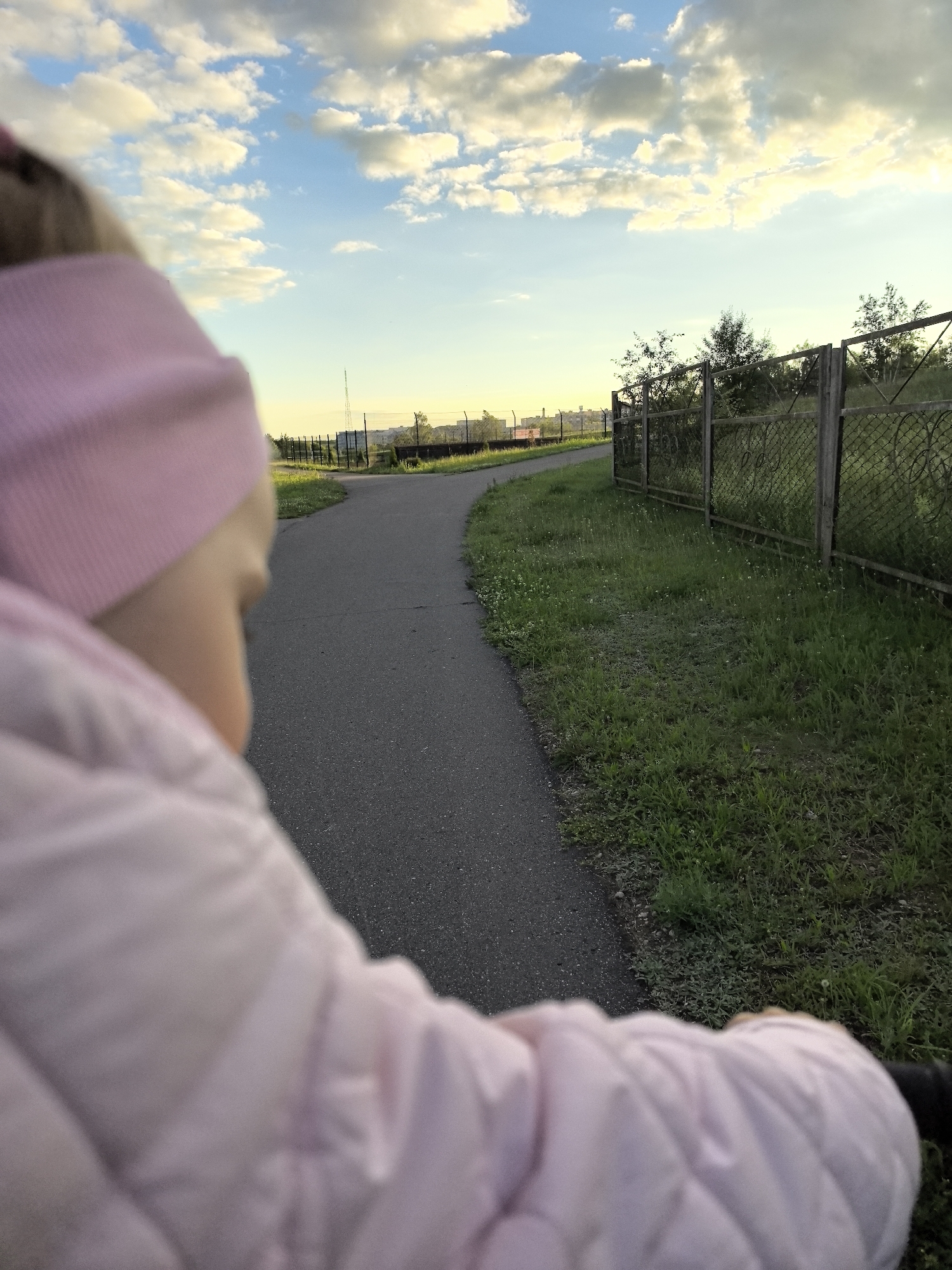 An individual in a pink beanie and a pink puffer jacket is shown from behind, looking towards a paved pathway that curves to the left. The pathway is bordered by a grassy area and a metal fence with a diamond pattern. In the background, there are trees, distant structures, and a tall antenna. The sky is blue with scattered white and yellow clouds, suggesting it is either sunrise or sunset. The location is described as Unknown, Unknown.