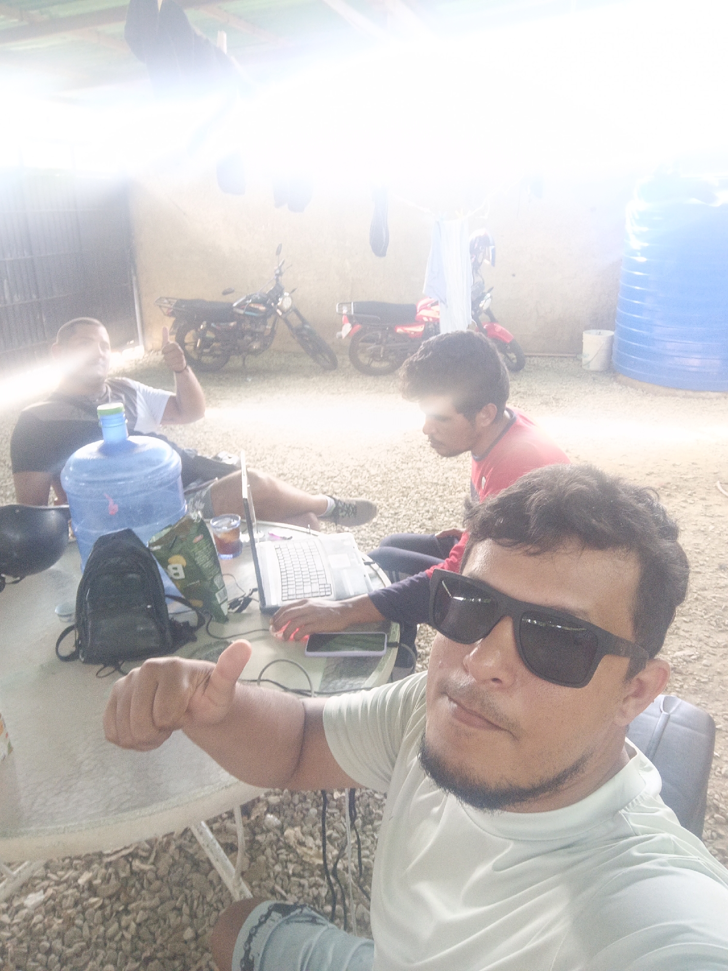 In Juan Griego, Venezuela, three individuals are gathered around a table outdoors, possibly under a covered area. A person in the foreground, wearing sunglasses and a light-colored shirt, is taking a selfie with a thumb up. To their left, a large blue water jug and a black backpack sit on the table. A laptop is open, and a hand with illuminated red fingertips is interacting with it. In the background, another person wearing a red shirt is seated. Further back, a third individual, wearing a black and white shirt, is also seated, with a thumbs-up gesture. Two motorcycles are visible behind the seated individuals. The ground is covered in gravel, and a large blue water tank stands to the right of the frame. The scene suggests a casual, outdoor workspace or gathering.