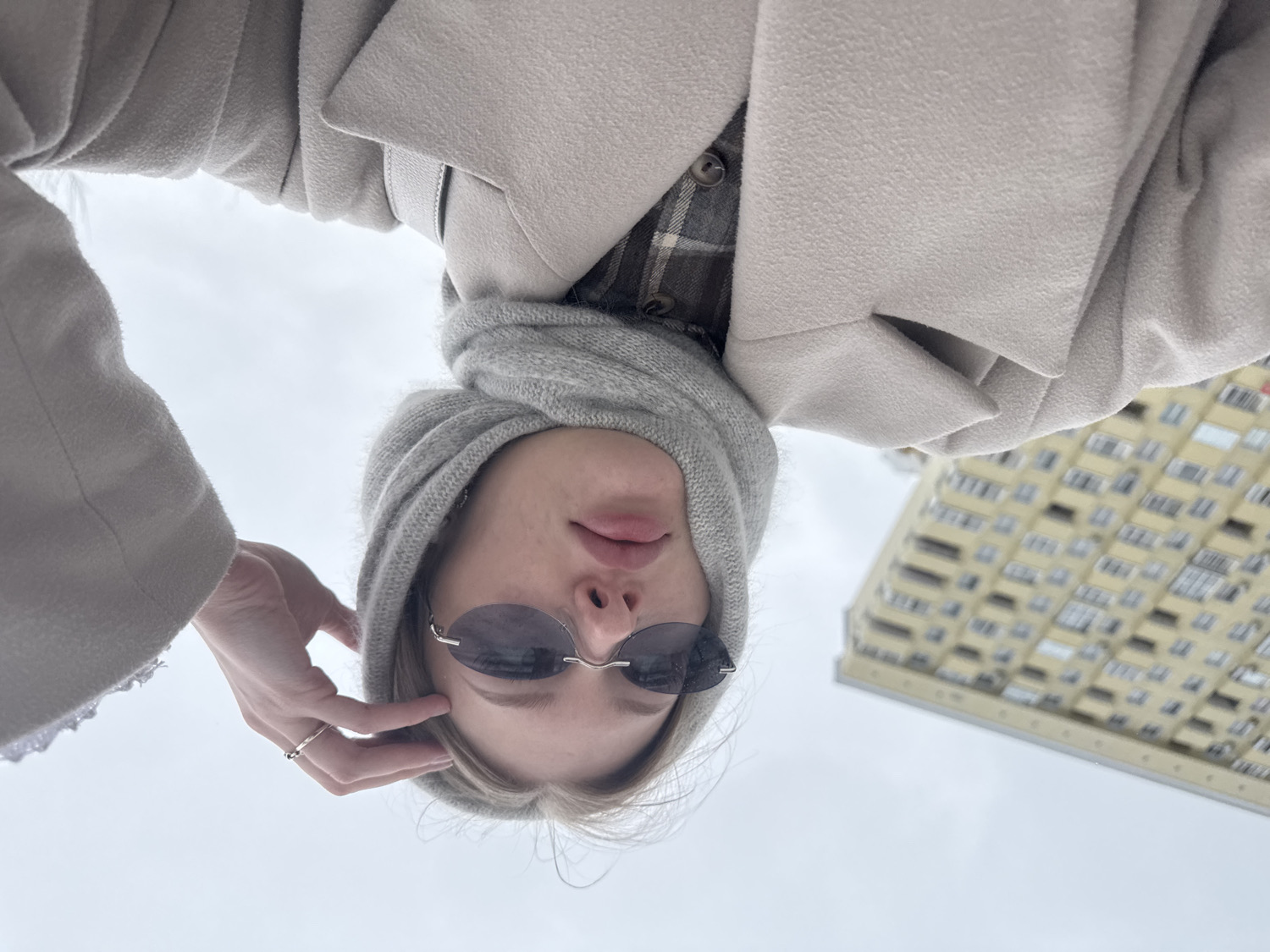 An upside-down, low-angle shot depicts a person looking downwards, seemingly taking a selfie. The person is dressed in winter attire, including a light beige overcoat worn over a plaid shirt. A grey knitted hood or scarf covers their head and neck, framing their face. They wear small, rimless sunglasses with dark lenses. Their lips are slightly parted. The person's left hand is raised to their temple, revealing a simple ring on their ring finger. The background is predominantly a bright, overcast sky, suggesting a cloudy day. To the right, a tall, multi-story building with numerous windows, light yellow or beige in color, is visible, tilting into the frame due to the extreme low-angle perspective. The overall scene conveys a casual outdoor moment in what appears to be cold weather, consistent with the location context of Perm, Russia. There is no visible text in the image.
