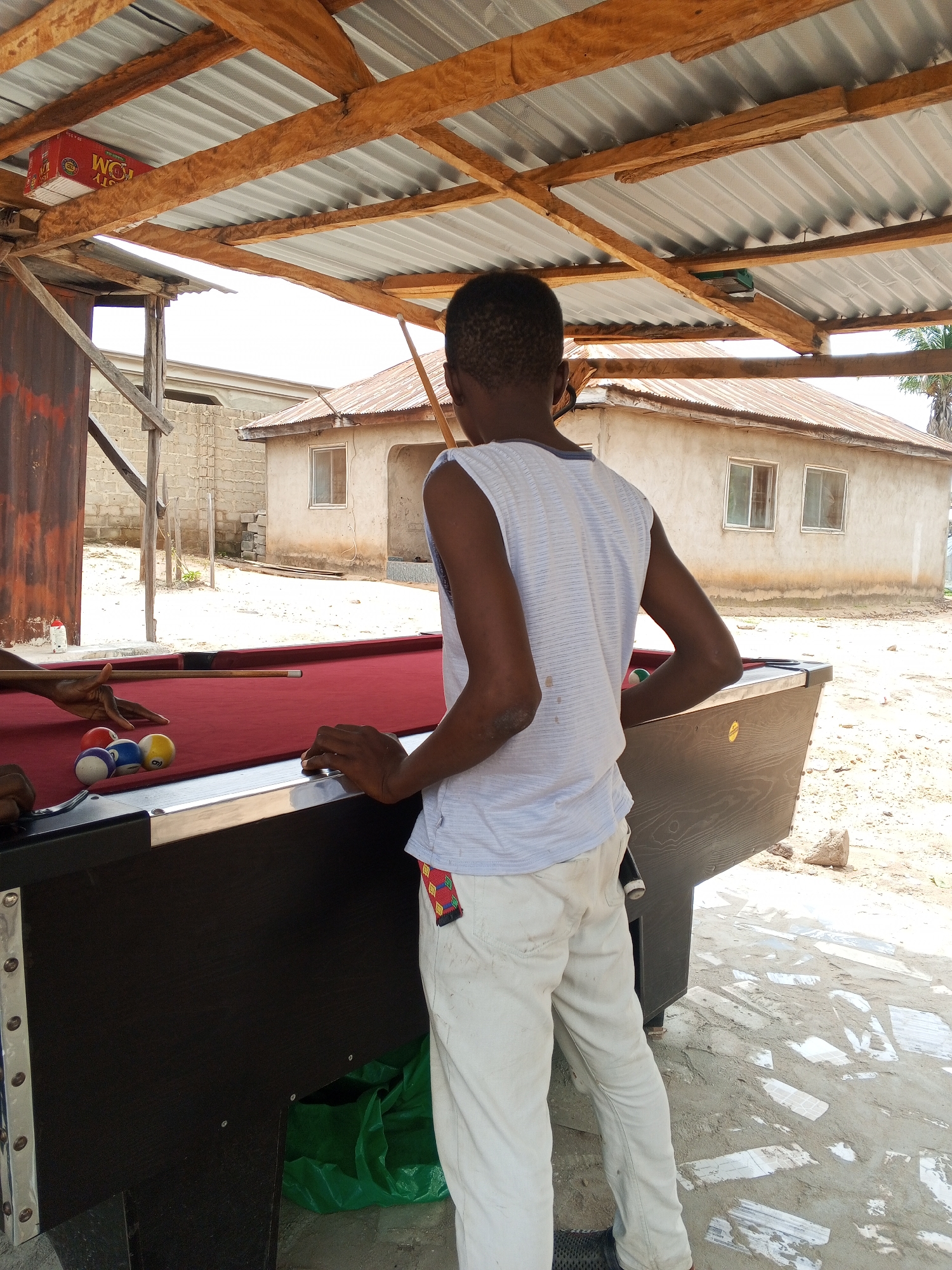 In the city of Lokoja, Nigeria, under a corrugated metal roof supported by wooden beams, a young person stands by a pool table. The individual, wearing a white and blue striped sleeveless shirt and off-white pants with a colorful belt detail, is positioned behind the table, facing away from the camera. The pool table, with a red felt surface, is partially visible, and several pool balls, including a red, a blue, and a yellow numbered ball, are scattered on the felt. Another person's hands are visible on the left side of the table, holding a pool cue and appearing to be in the process of playing. The environment is outdoors, with sandy ground and some scattered stones. In the background, a simple building with a tin roof and windows can be seen. A red box with white lettering, possibly a product container, is affixed to the wooden beams overhead. A green plastic bag is on the ground near the pool table.