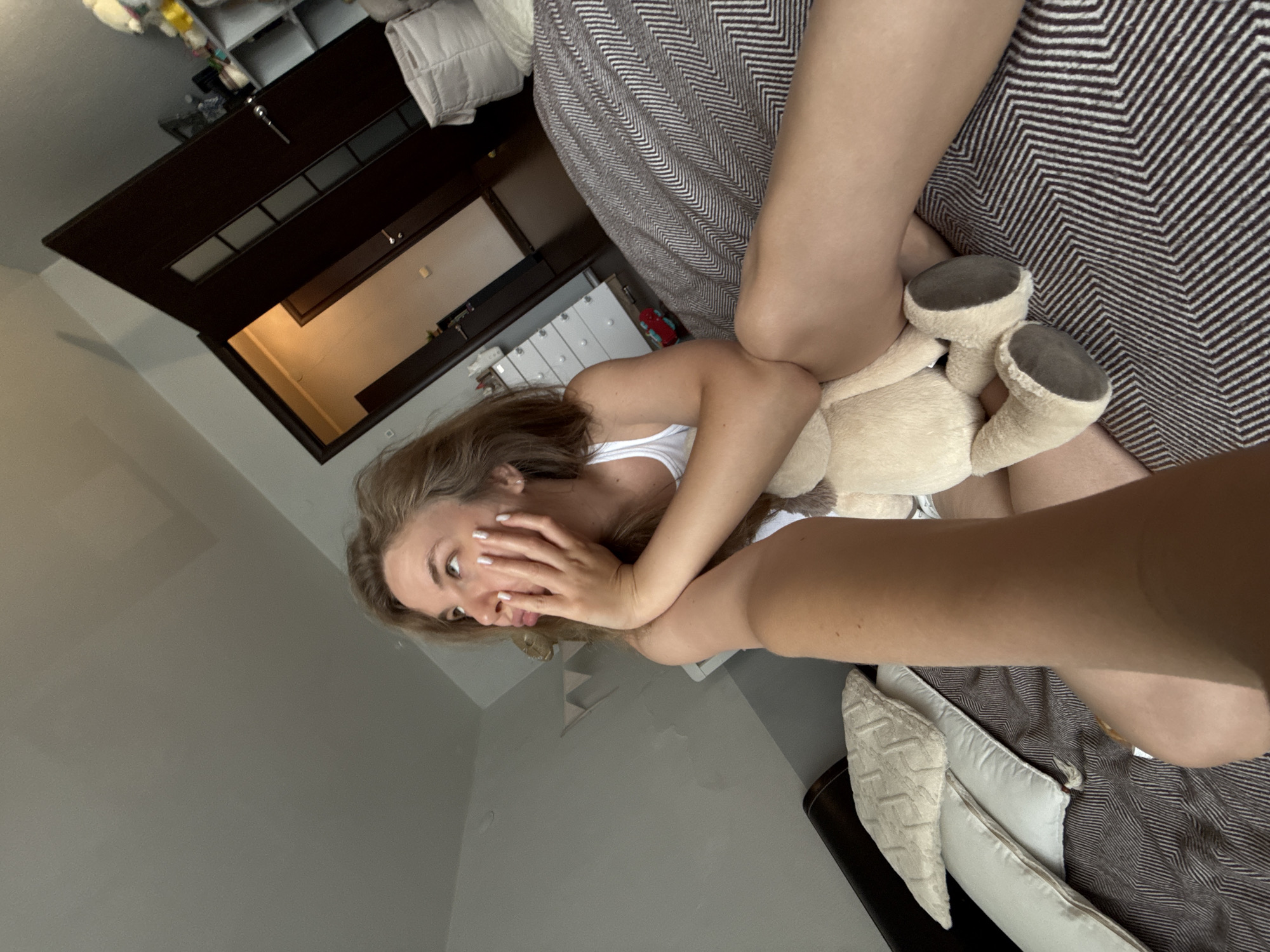 The image shows a young woman in a white tank top lying on a bed with a stuffed animal. She is resting her head on her hand and looking up and to the side with a pouty expression. The room appears to be a bedroom with a dresser and a doorway visible in the background. There is a patterned blanket on the bed, and pillows are arranged near the headboard. The overall impression is that of a casual, indoor setting. The image does not contain any specific details that would indicate its location in Vladimir, Russia.
