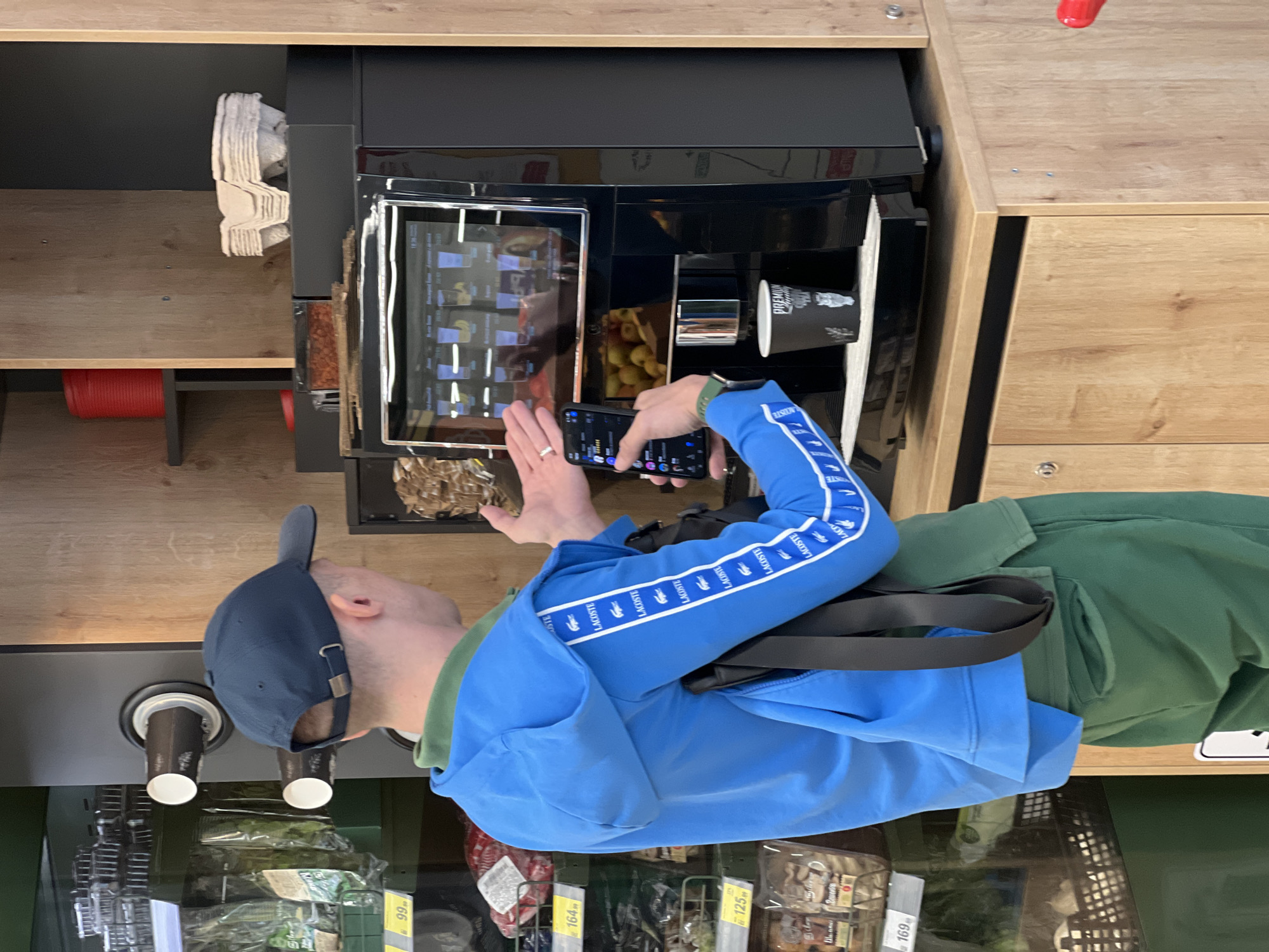 A person stands in front of a self-serve coffee machine in what appears to be a supermarket in Krasnodar, Russia. The person is wearing a blue baseball cap, a blue Lacoste-branded tracksuit, and a green t-shirt. A black shoulder bag is visible. They are holding a smartphone and seem to be taking a photo of the coffee machine's screen. The coffee machine is dispensing a drink into a paper cup. The surrounding environment includes a wooden cabinet that houses the coffee machine, and below it are shelves with various food products, including packaged meats and vegetables. Price tags in Russian currency (rubles) are visible on the shelves.