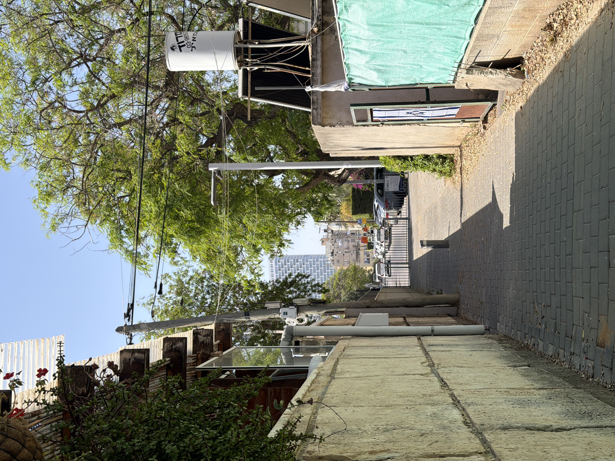 The image shows a narrow street in Bat Yam, Israel. The street is paved with gray brick. Buildings line both sides of the street. One building has a water tank on its roof and a green tarp hanging over the side. Another building has a corrugated metal fence. A tree with green leaves overhangs the street. In the distance, tall buildings are visible. Cars and scooters are parked along the street. Overhead power lines are strung between poles. The sky is blue.