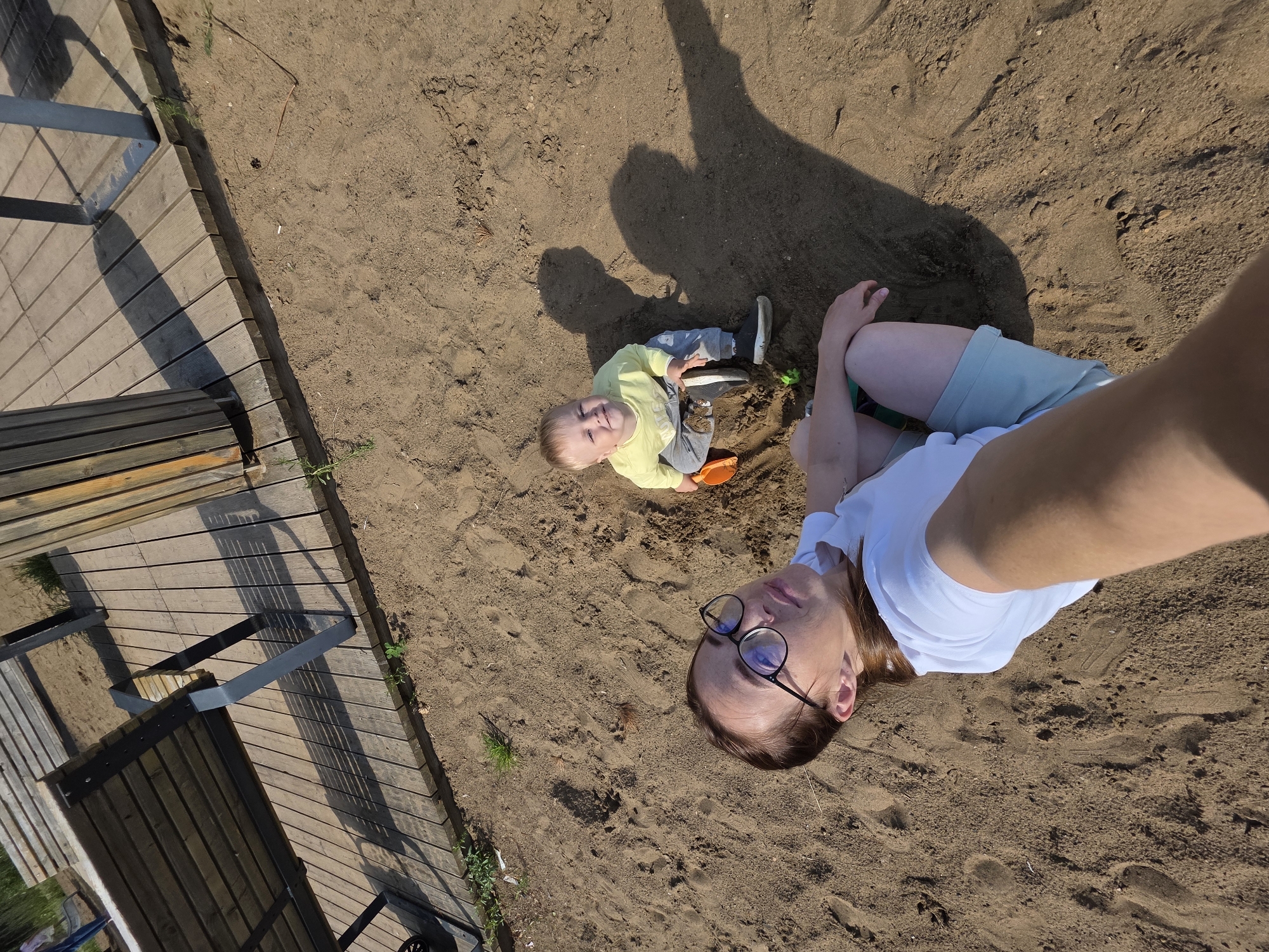 This high-angle, full-shot photograph captures a mother and child on a sandy area, likely near a wooden boardwalk structure, possibly in Chaikovsky, Russia. The mother, in the foreground, is lying on her back, taking a selfie with her arm extended. She wears a white t-shirt and light blue shorts, with black-rimmed glasses. To her right, a young child, around two years old, is sitting in the sand, wearing a yellow long-sleeved shirt and gray pants. The child is holding an orange toy shovel and appears to be playing in the sand. The environment consists of sand with footprints and scattered debris like small twigs. Behind them, a wooden platform with planks and benches is visible, casting long shadows due to the sunlight. The overall scene suggests a casual outdoor activity in a park or recreational area.