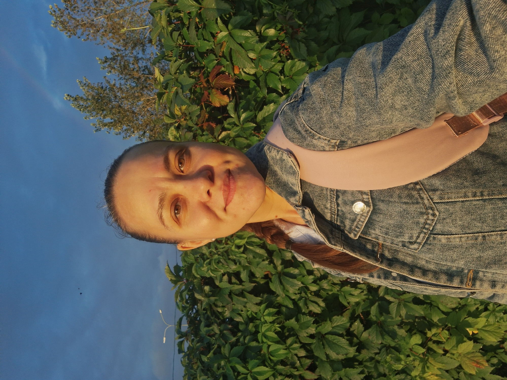 In Chaikovsky, Russia, a young woman with brown hair braided down her back takes a selfie in front of a dense green hedge. She is wearing a denim jacket over a light-colored shirt and a light pink backpack. The sky above is a deep blue with a hint of a rainbow on the right side. Streetlights are visible in the distance to the left. The lighting suggests it is late afternoon or early evening, with the sun casting a warm glow on the woman's face.