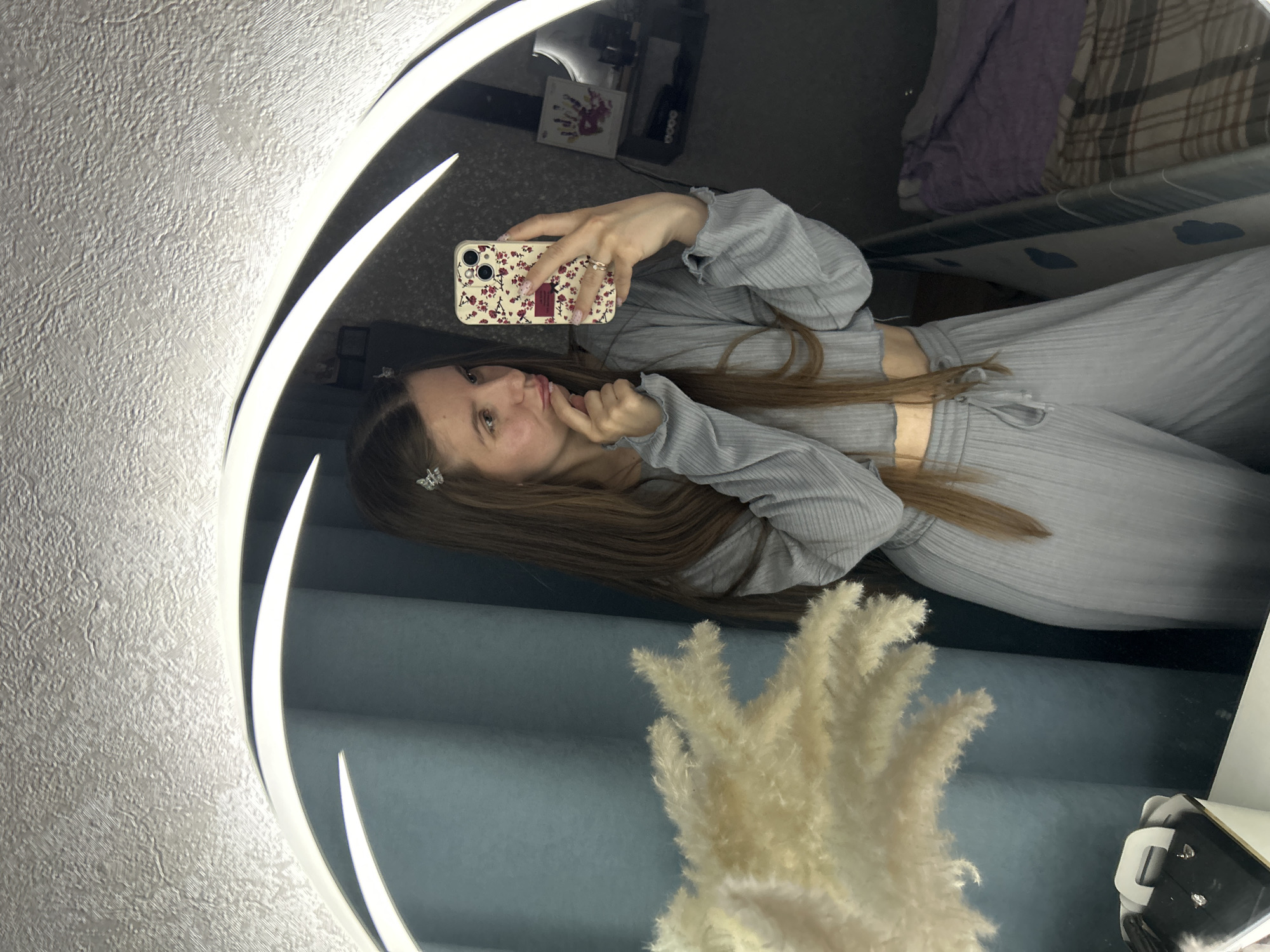 The image displays a young woman taking a mirror selfie in an indoor setting. The photo is rotated 90 degrees counter-clockwise, making the woman appear to be positioned horizontally. She has long, straight brown hair and is wearing a light gray ribbed two-piece outfit, comprising a crop top and matching pants. A small, silvery butterfly hair clip is visible in her hair. Her fingernails have a light-colored polish, and she wears a ring on her left ring finger.
She holds a smartphone in her right hand, capturing her reflection. The phone is adorned with a light-colored case featuring a patterned design of small red flowers and black accents. Her expression is a playful pout, with her left index finger gently touching her lips.
The mirror itself is round and features a prominent, illuminated white light strip along its inner edge, providing bright, direct lighting.
Reflected in the mirror, behind the woman, is a blue or teal-colored wall. Further back, in the upper right quadrant of the reflection, part of a bed with purple and plaid bedding can be seen, indicating a bedroom or living space. Other reflections include a small, framed item resembling a child's handprint and some dark electronic equipment (possibly a camera or lighting setup).
In the lower right section of the mirror's reflection, a large, fluffy, light-colored decorative plant, likely pampas grass, is visible. The wall adjacent to the mirror (on the viewer's left) features a textured, light gray or white wallpaper. A small section of an electronic device or charger is also visible in the bottom right corner of the reflected scene. No discernible text is visible in the image.