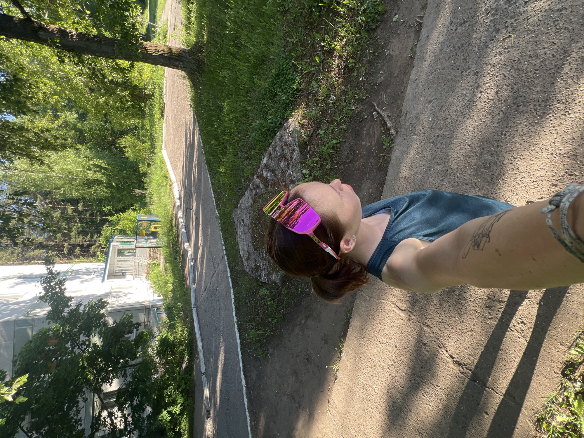 The image shows a woman taking a selfie in an outdoor environment in Chaikovsky, Russia. She's wearing a dark blue tank top and bright pink sunglasses. Her hair is pulled back in a ponytail. The location appears to be a sidewalk or paved path adjacent to a grassy area with trees and a building in the background. The building is light-colored and seems to be a multi-story structure. The pathway is made of concrete and has visible cracks. Shadows cast by trees and other objects are apparent on the pavement. A small area with grass and shrubbery is seen between the walkway and the building.
