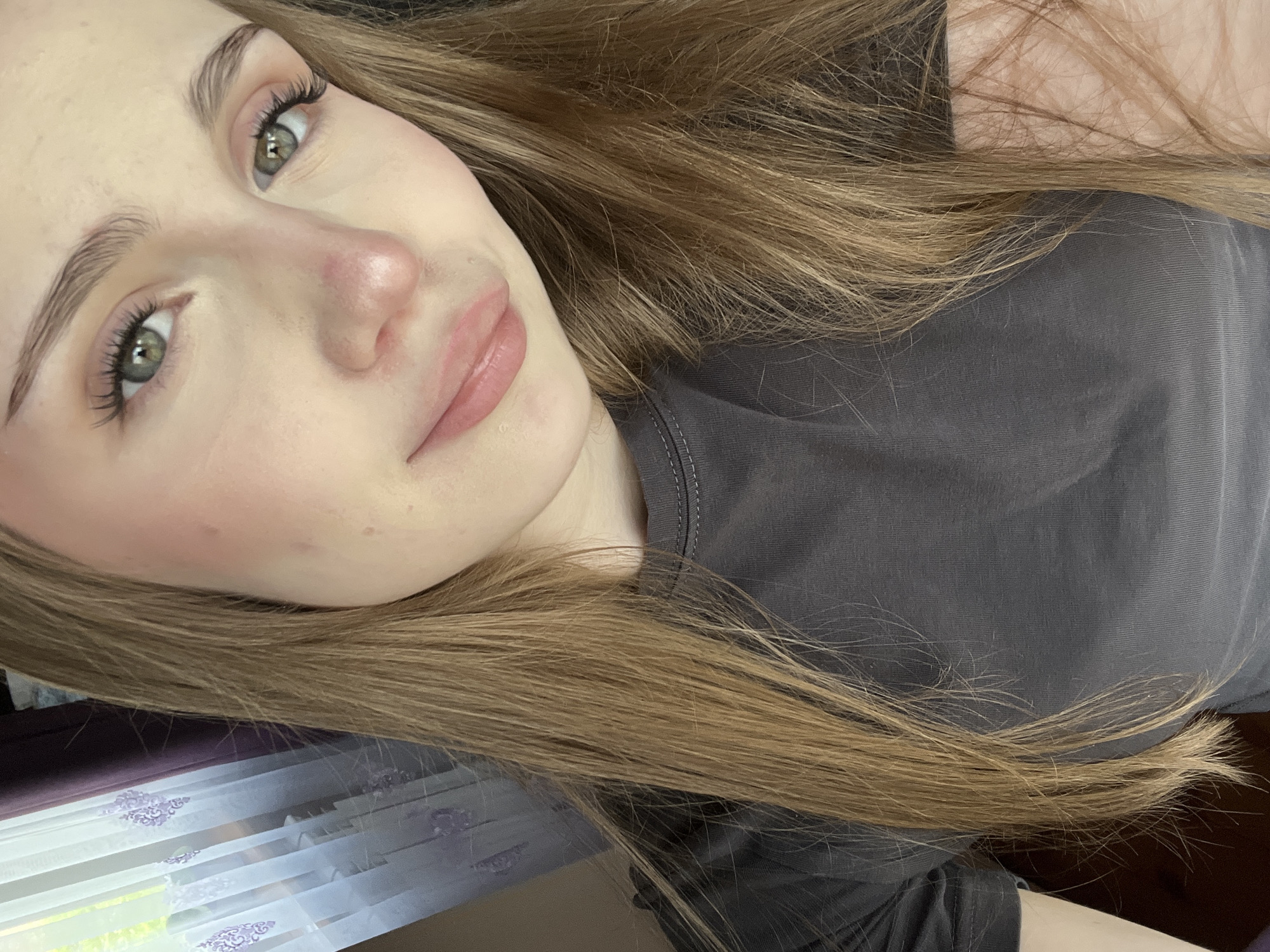 A close-up, waist-up, indoor portrait shows a young woman with long, straight, light brown hair. She is wearing a dark grey t-shirt with a ribbed neckline. Her skin is pale with a light blush on her cheeks. She has green eyes, light brown eyebrows, and light pink lips. Her hair is parted on the side and falls over her shoulders. The background includes a blurred window with sheer white curtains featuring a purple floral pattern. There is also a hint of a dark purple curtain to the left of the window. The lighting is natural and diffused, suggesting it may be daytime. The location is not specified to be in Ashkadar, Russia, as this information is not present in the image.