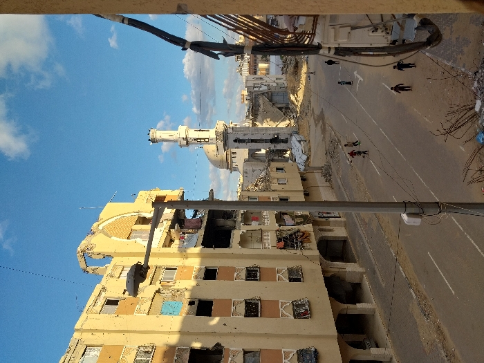 The image depicts a street scene in Khan Yunis, Palestinian Territory, characterized by significant urban destruction. In the foreground, a gray street light pole stands slightly right of center, with utility wires extending across the frame. To the left, a multi-story residential building, light tan in color, shows extensive structural damage, including large sections of its upper floors and facade missing, revealing interior spaces. Multiple window openings are empty, and some balconies display hanging laundry. On the right side of the street, the immediate foreground contains rubble, twisted rebar, and debris piles.
Further down the street, centrally located, is a paved road with visible white lane markings. Approximately seven individuals, appearing to be mostly children, are present on the road. Three children on the left side of the road appear to be walking or playing, one possibly holding a ball. Three or four other figures are further down the street on the right.
In the mid-ground and background, more damaged buildings line the street. A prominent mosque structure is visible, featuring a largely intact white minaret and a partially damaged dome, with surrounding structures reduced to rubble. The area around the mosque also exhibits significant destruction. The sky above is blue with scattered white clouds, and sunlight casts shadows, indicating daytime. Utility cables are visible on the far right edge of the frame.