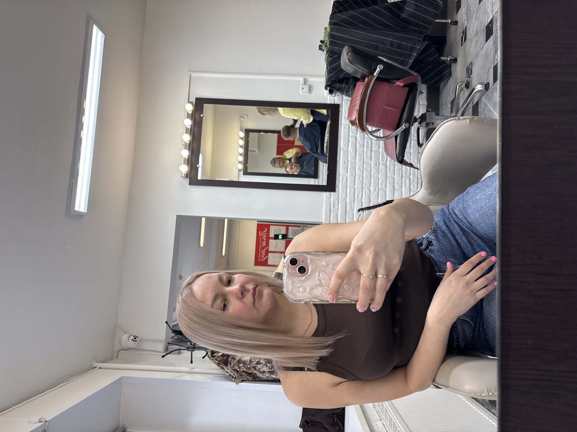 A young woman sits in a salon chair, taking a selfie. She has blonde hair and is wearing a brown tank top and jeans. She is holding a phone with a light pink case. A mirror behind her reflects other people in the salon. The salon's interior is visible, with white walls, bright lights, and styling stations. Red and black bags are on one of the chairs and the location is Izhevsk, Russia.