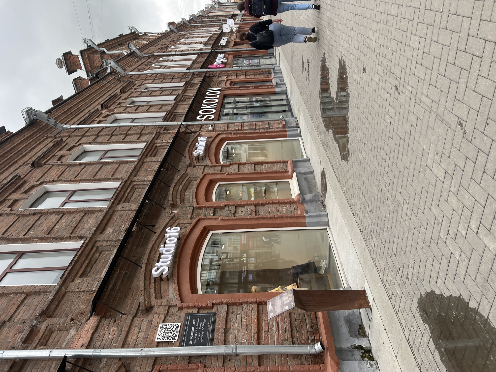 The image depicts a street scene in Yekaterinburg, Russia, on a cloudy day. A two-story brick building with arched windows and signage for businesses lines the right side of the frame. Prominent signs include "Studio16" and "SOKOLOV". The building has a classic architectural style with decorative brickwork and metal gutters.
In front of the building is a paved sidewalk with a herringbone pattern. Several puddles of water are visible on the sidewalk, reflecting the building and the sky.
Two women are walking away from the camera on the sidewalk. The woman on the left is wearing a black backpack and blue jeans. The woman on the right is wearing a dark backpack and blue jeans.
To the left of the building, a metal downspout is visible, running vertically along the brick facade. A QR code and an informational plaque are mounted on the brick wall near the downspout, indicating historical information about the building. A wooden easel with information is placed on the sidewalk.
The overall impression is a glimpse of a historical urban environment in Yekaterinburg.