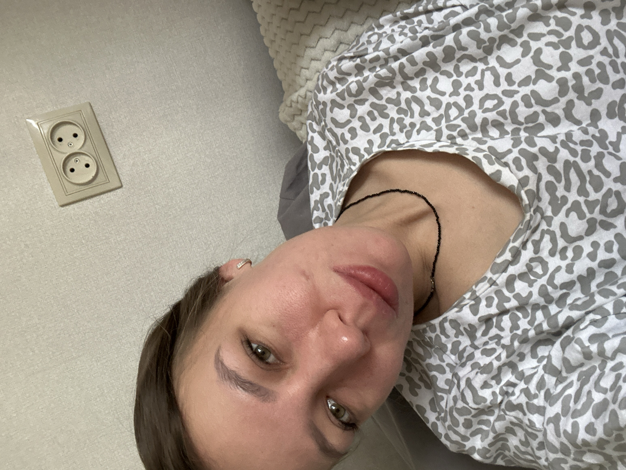 A woman with brown hair and green/hazel eyes is prominently featured in this close-up indoor selfie, looking directly at the camera with a neutral expression. She has full, glossy lips and wears a delicate dark beaded necklace and a small hoop earring in her right ear. Her skin appears slightly flushed.
She is wearing a light-colored top with a gray leopard-spot pattern. A soft, textured, light gray fabric, possibly a blanket or pillow, is visible behind her head and shoulder, suggesting she is lying down or reclining.
To the left of the woman, mounted on a light gray, subtly textured wall, is a double electrical outlet. The outlet design, with two round sockets and a grounding pin hole in the upper socket, is consistent with Type F (Schuko) sockets commonly used in Tomilino, Russia, aligning with the provided location context. The overall setting appears to be a domestic indoor space, likely a bedroom or living area. No discernible text is visible in the image.