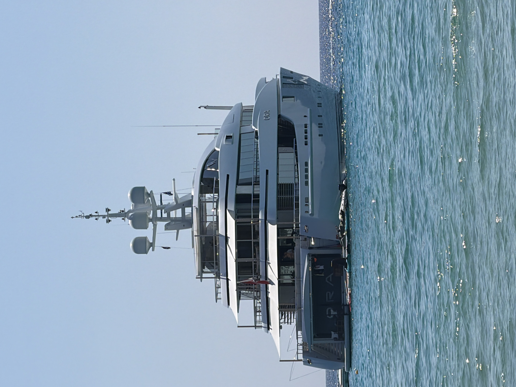 A large, white luxury yacht named "ORA" is anchored in the blue, sun-dappled water of the Aegean Sea, under a clear sky. The yacht has multiple decks, large windows, and a prominent radar and communication mast. The stern of the yacht features an open garage area with what appears to be some water sports equipment. There are a few people visible on the yacht's decks. The water is calm with small waves reflecting the sunlight. The location is not specified within Greece, but the scene suggests a popular coastal or island destination.