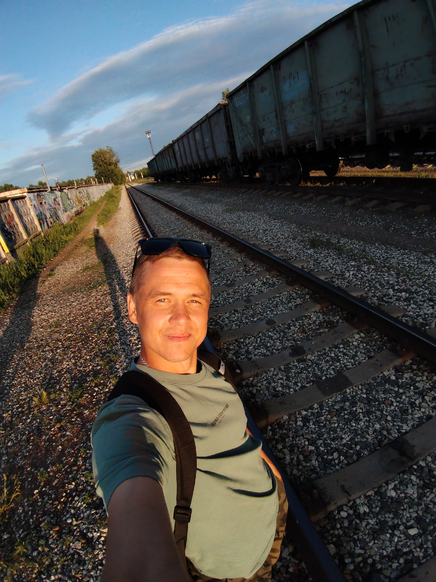 A male individual is depicted in a selfie taken outdoors near railway tracks in Chaikovsky, Russia. The subject, appearing to be an adult male with short brown hair, fair skin, and blue eyes, has sunglasses resting on top of his head and a silver chain around his neck. He is wearing a light green t-shirt with a faint "ICONS COUTURE" logo on the right chest area and a black backpack. He is standing on a gravel path adjacent to two parallel railway tracks. To the right, a line of stationary, weathered, dark grey freight cars (specifically open-top gondolas with visible numbers like "7112774" and "70") occupies one of the tracks. To the left, a concrete wall extends parallel to the tracks, featuring various colorful graffiti or murals. Sparse green vegetation grows between the gravel path and the wall. In the background, a deciduous tree is visible behind the wall, and a tall metal utility pole stands near the tracks. The sky overhead is clear blue with several elongated, light grey clouds. The scene is illuminated by golden hour light, casting long shadows from the individual and objects on the ground.