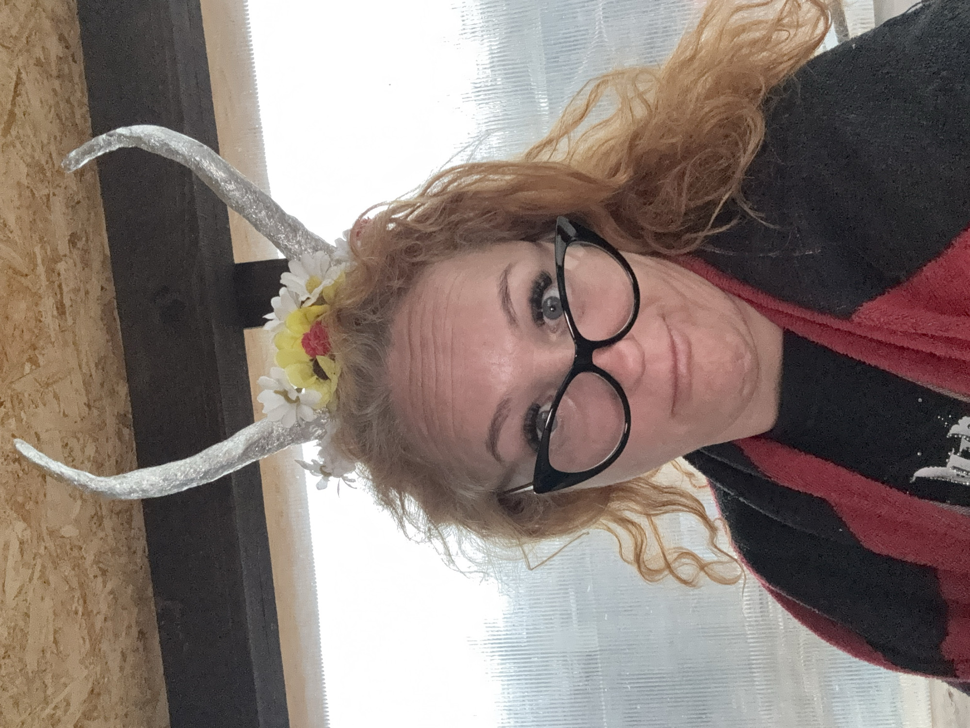 A woman with red curly hair and black-framed glasses is wearing a headband with silver horns and flowers. She is looking directly at the camera. She wears a red and black striped sweater. The background appears to be an interior with translucent corrugated plastic panels. The location is unknown and there is no information to determine if this is in Chaikovsky, Russia.