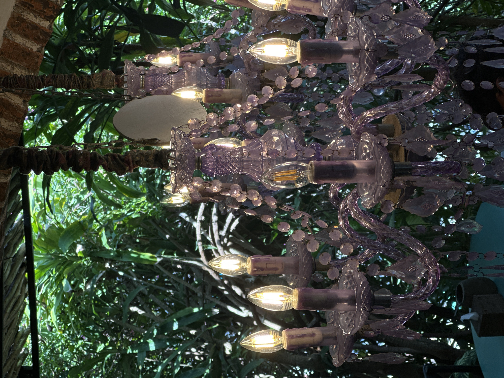 The image depicts a chandelier with purple crystal ornaments and several lit candelabra-style bulbs, suspended in an outdoor setting likely in Jimbaran, Indonesia. Lush greenery forms the backdrop, visible through the chandelier's structure. A portion of what appears to be a brick arch is visible on the left side of the image, indicating an architectural element.