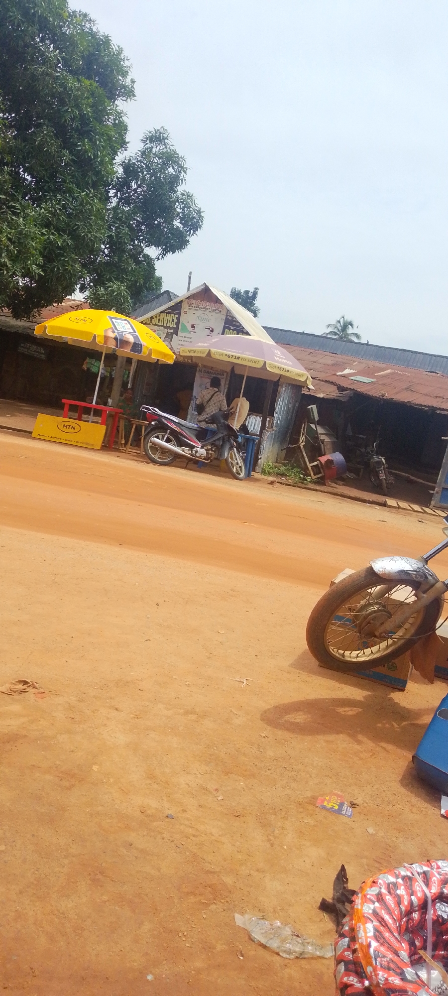 This image depicts a roadside scene in Baissa, Nigeria, on a bright, sunny day. The foreground is dominated by a dusty, orange dirt road, with a motorcycle parked on the right side, its front wheel and handlebars prominently visible. Beyond the road, a row of small shops or stalls is lined up, suggesting a marketplace or commercial area.
Several key objects stand out: two large, colorful umbrellas, one bright yellow with "MTN" branding, and another purple one, are set up in front of the shops, providing shade. A motorcycle is parked under the purple umbrella, and a person is seated on it, partially obscured. A red table with "MTN" branding is positioned near the yellow umbrella. Various signs are visible on the shops, including one that reads "SERVICE" and others with "MTN" logos and phone numbers. A discarded plastic bottle and some other debris are scattered on the road. In the background, lush green trees frame the scene, indicating a natural environment surrounding the settlement. The overall impression is that of a typical day at a bustling roadside commercial hub in a rural or semi-urban area of Nigeria.