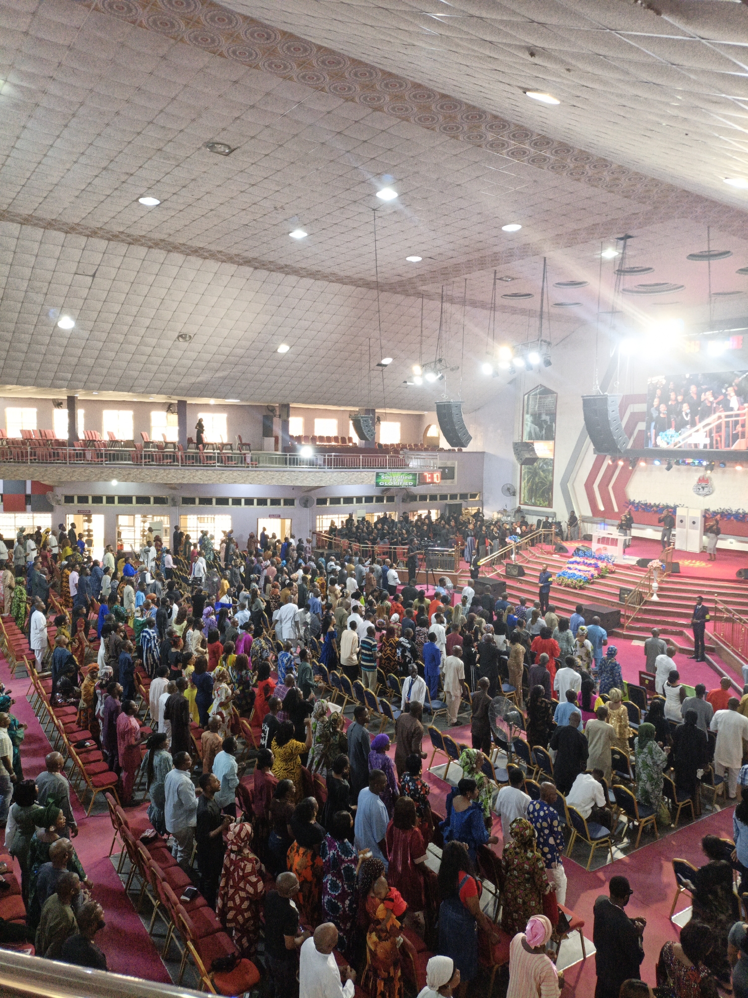 Here's a detailed description of the image:
**Location and Environment:**
The image appears to be taken inside a large church or auditorium in Sabon Gari West, Nigeria. The architecture includes a high, patterned ceiling with recessed lighting, a balcony area overlooking the main floor, and a prominent stage area.
**People:**
The space is filled with a large crowd of people, primarily adults. They are dressed in a variety of clothing, many in colorful and traditional African attire. Some individuals are standing, while others are seated in rows of chairs. Some individuals are standing on the balcony.
**Objects:**
Visible objects include rows of chairs, speakers hanging from the ceiling, a large screen on the stage, and various decorations on the stage itself (including floral arrangements and coverings).
**Actions/Interactions:**
The people are generally facing the stage area. It is likely that they are participating in a church service, conference, or similar event.