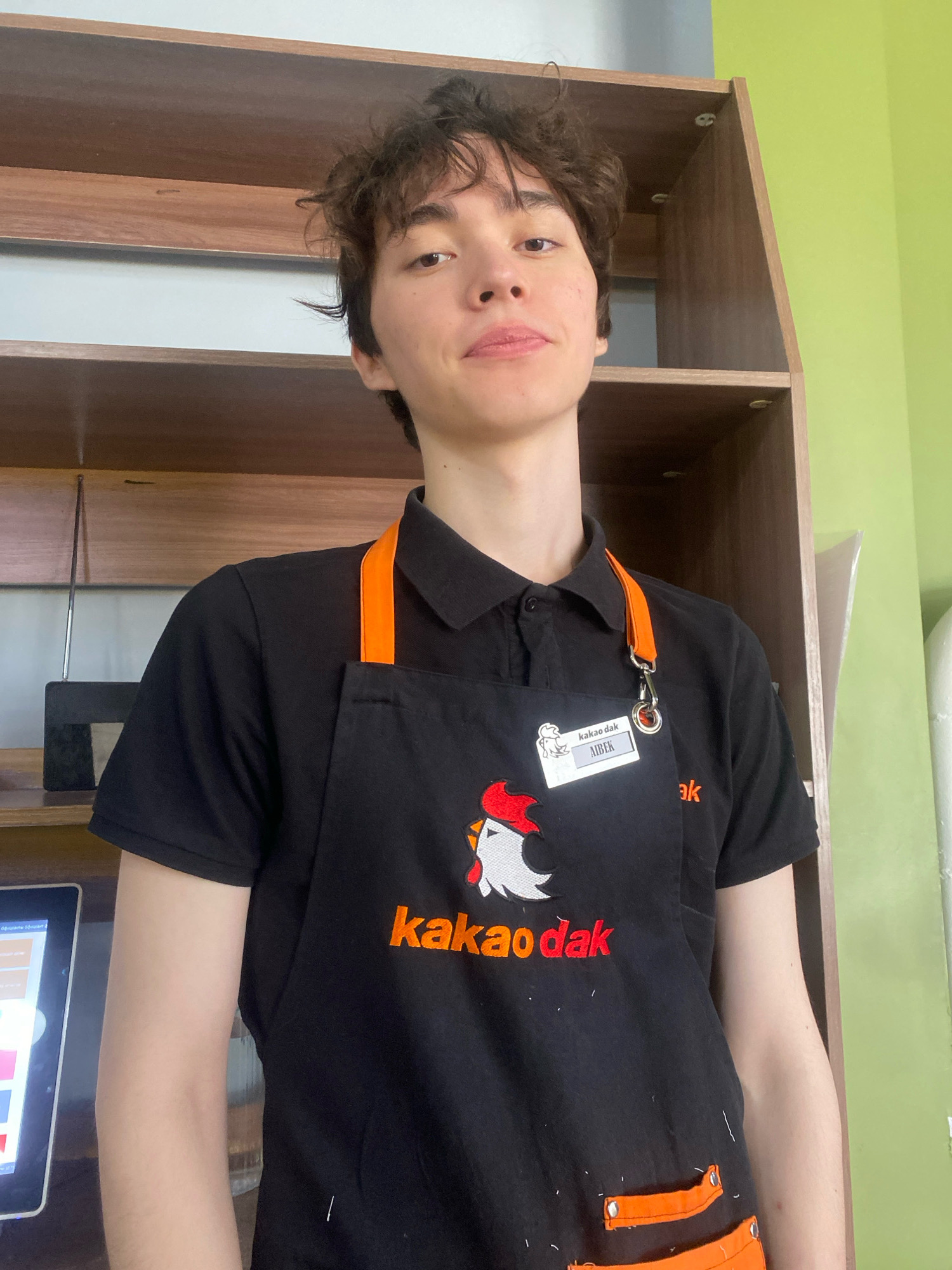 A young person with dark, wavy hair is standing and looking at the camera. They are wearing a black polo shirt with an orange-trimmed black apron over it. The apron has a logo of a rooster and the text "kakao dak" in orange lettering. A name tag on the apron reads "AIbEK" in white text on a grey background. The person is standing in front of a wooden shelf unit with shelves of varying heights. To the right of the shelf unit is a lime green wall. In the lower left corner of the image, a tablet device is partially visible, displaying colored squares and text. The overall environment appears to be an indoor commercial space, possibly a food establishment, in Astana, Kazakhstan.