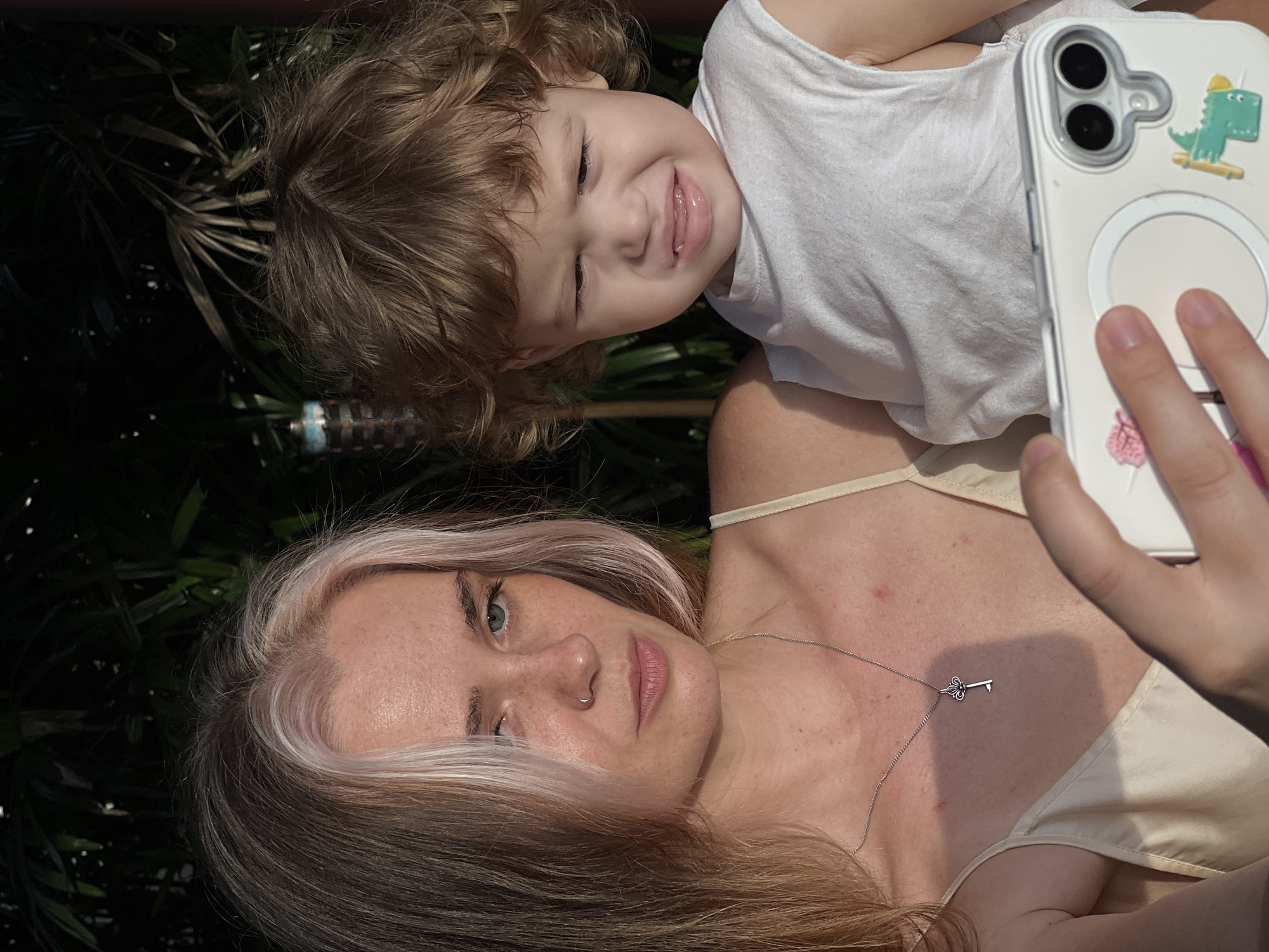 A close-up, outdoor, waist-up shot captures a woman holding a toddler. The woman, on the left, has blonde hair with bleached streaks and is wearing a light-colored camisole. She is looking down and to the left, seemingly at a phone held by her right hand. A silver key-shaped pendant hangs from a chain around her neck. The toddler, on the right, is looking towards the woman with a distressed facial expression, its mouth agape and eyes squinted. The toddler wears a white sleeveless shirt. The background consists of dark foliage and a torch-like object, suggesting an outdoor setting, possibly a garden or patio. The lighting suggests it is late afternoon or early evening.