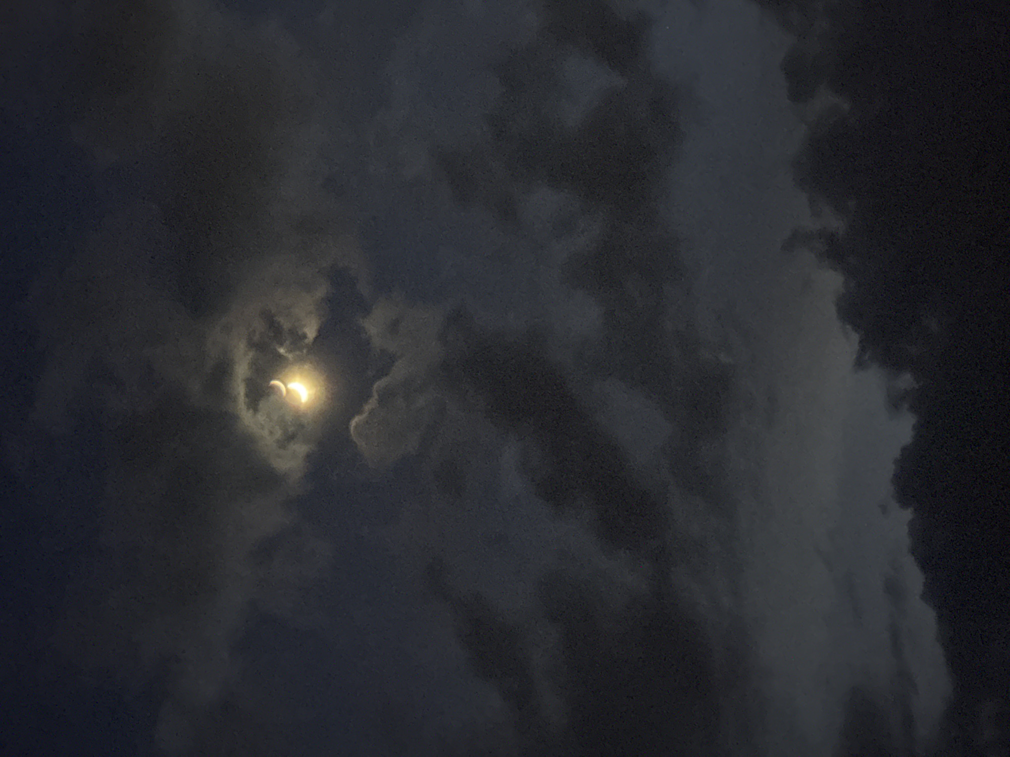 The image shows a partially obscured view of the sky at night, with a bright celestial object, potentially the moon or a planet, shining through a gap in the clouds. The sky is filled with dark, layered clouds in varying shades of gray and black. The brightest point is at the upper left, where the source of light is visible. The environment is dark and cloudy, suggesting a potentially stormy or overcast night. The location in the city of Pleven, Bulgaria is not discernible from this image. There are no people or identifiable objects present in the frame other than the sky and clouds.