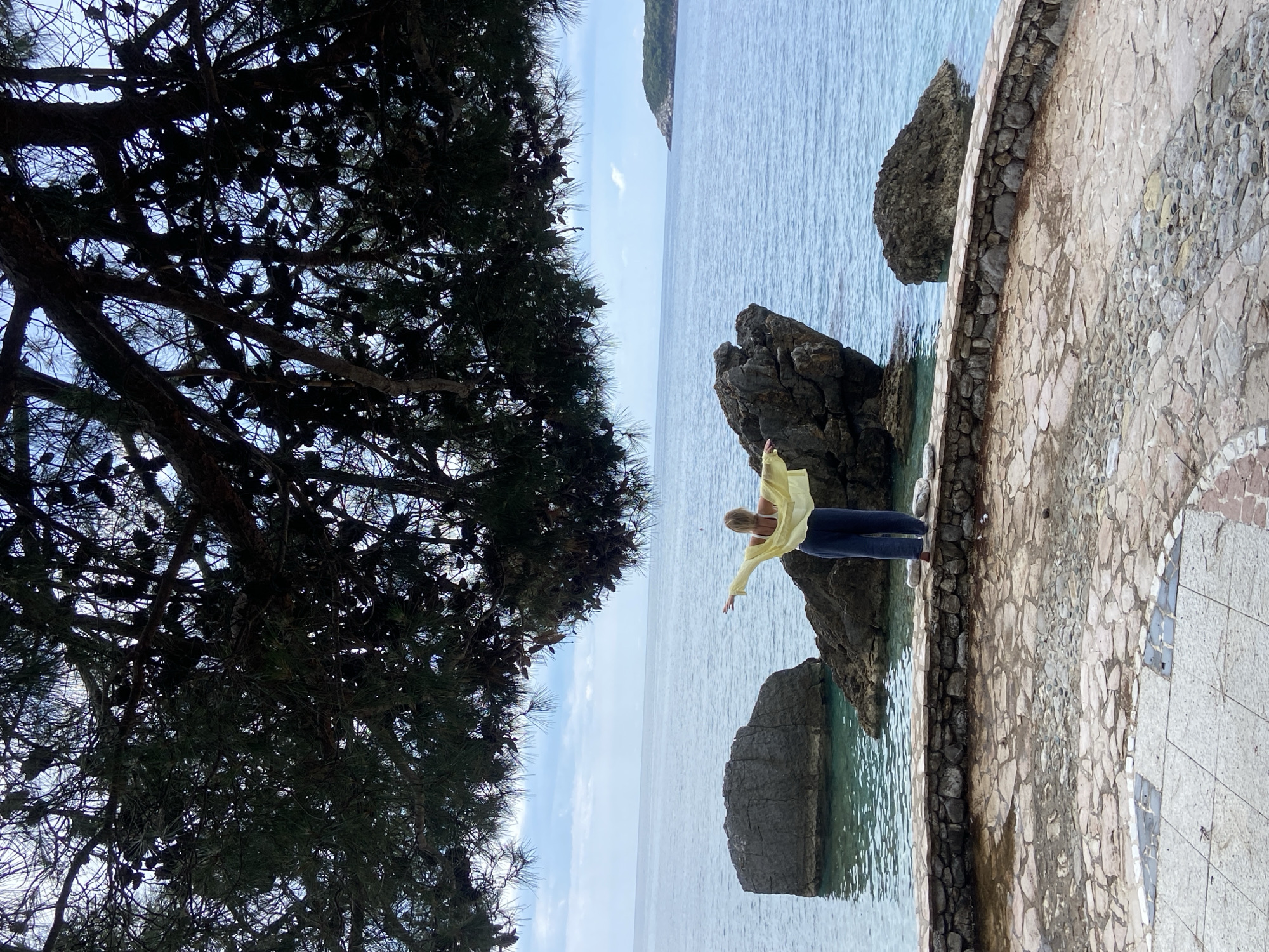 A medium shot shows a blonde woman with outstretched arms, standing on a stone ledge next to the Adriatic Sea in Prijevor, Montenegro. She wears a light yellow shirt and dark denim jeans. Behind her, large, dark rocks rise from the clear, turquoise water. To the left, a large evergreen tree with dark branches dominates the view. The background features a clear blue sky with a small island visible in the distance. The ledge consists of various light-colored stones.
